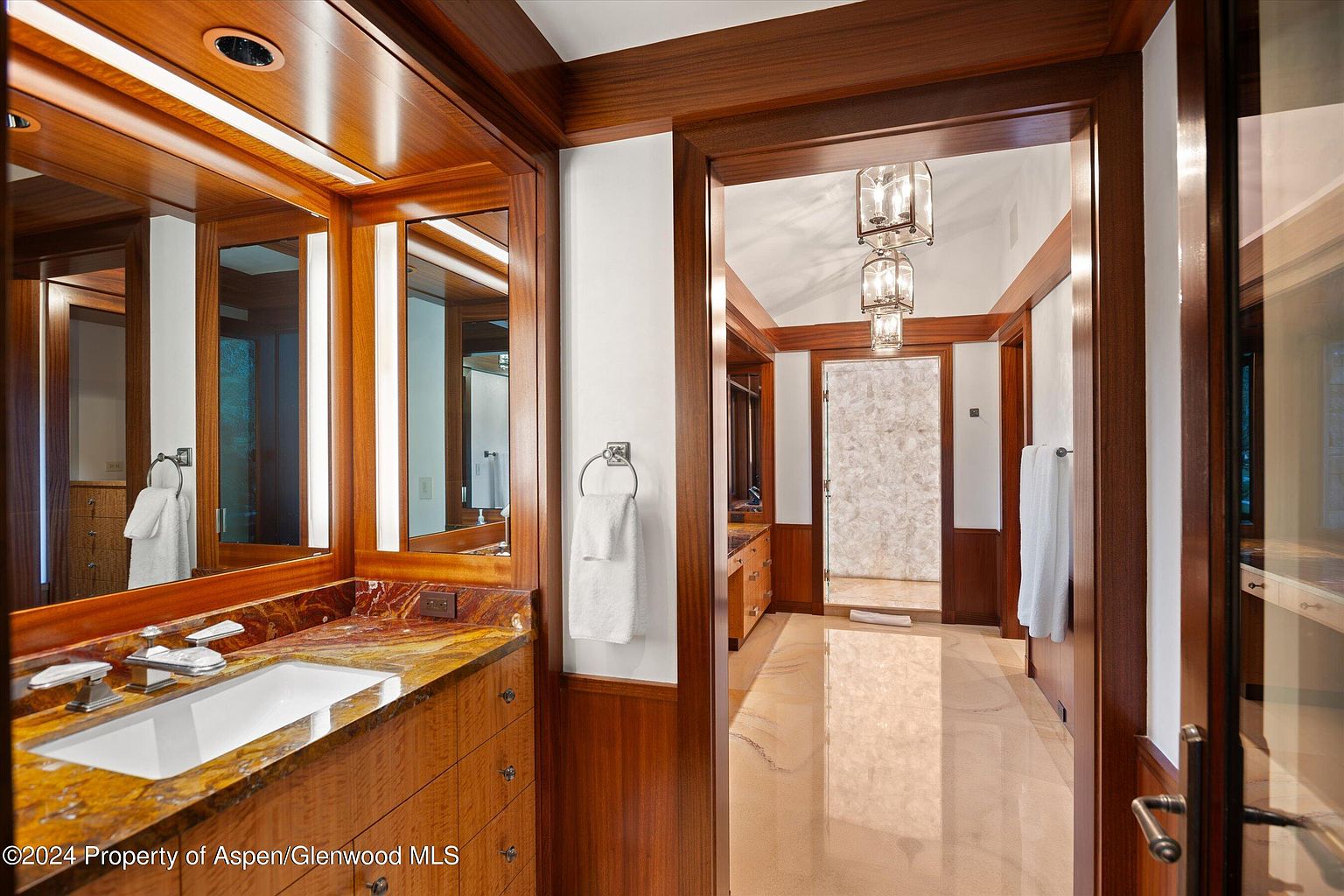 This is a well-appointed primary bathroom featuring rich wood cabinetry and trim, creating a warm and luxurious atmosphere. The vanity boasts a unique countertop with a vessel sink, complemented by a large mirror with integrated lighting. Through the doorway, a glimpse of the shower with decorative wall panels and elegant lighting fixtures adds to the sophisticated ambiance.