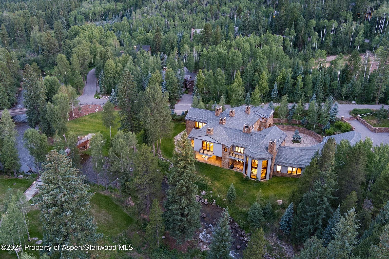 This aerial view showcases a luxurious home nestled amidst a dense forest, with a river flowing nearby. The house features a complex roofline, multiple chimneys, and large windows that suggest a well-lit interior. The surrounding landscape is lush and green, emphasizing the property's privacy and natural setting.