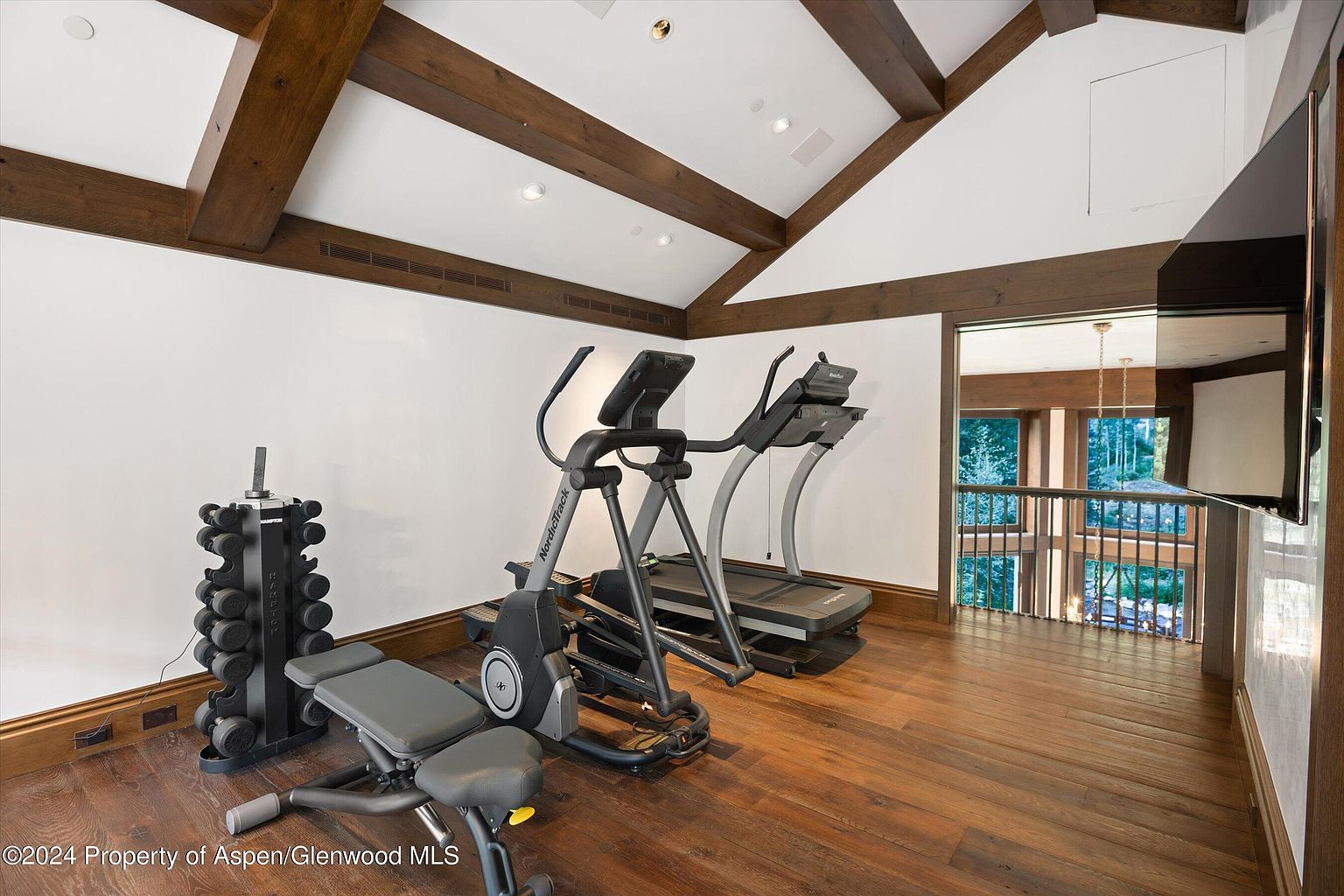 This is an interior shot of a home gym featuring hardwood floors, exposed wooden beams on the ceiling, and white walls. Exercise equipment such as a treadmill, elliptical machine, and weight rack are present. The room has a view into another area of the house, suggesting an open and airy layout.