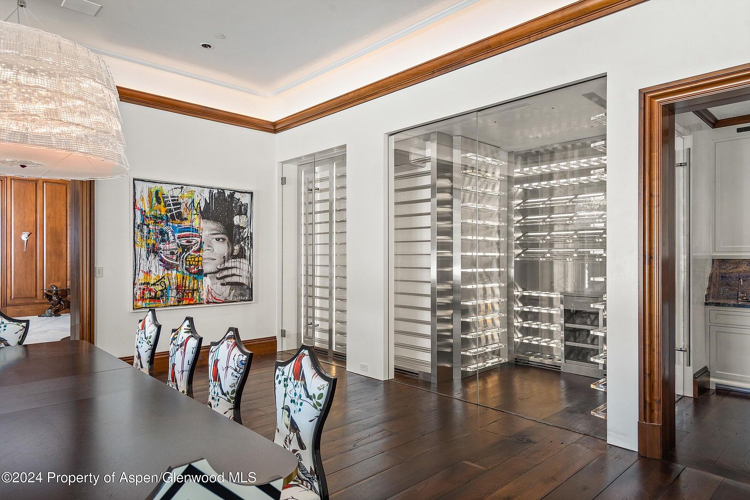 This interior shot showcases a sophisticated dining room with a large, dark wood table surrounded by elegantly patterned chairs. A vibrant, modern art piece hangs on the wall, adding a pop of color and contemporary flair. The room features a wine cellar with glass doors, highlighting the home's luxury amenities and creating a focal point.