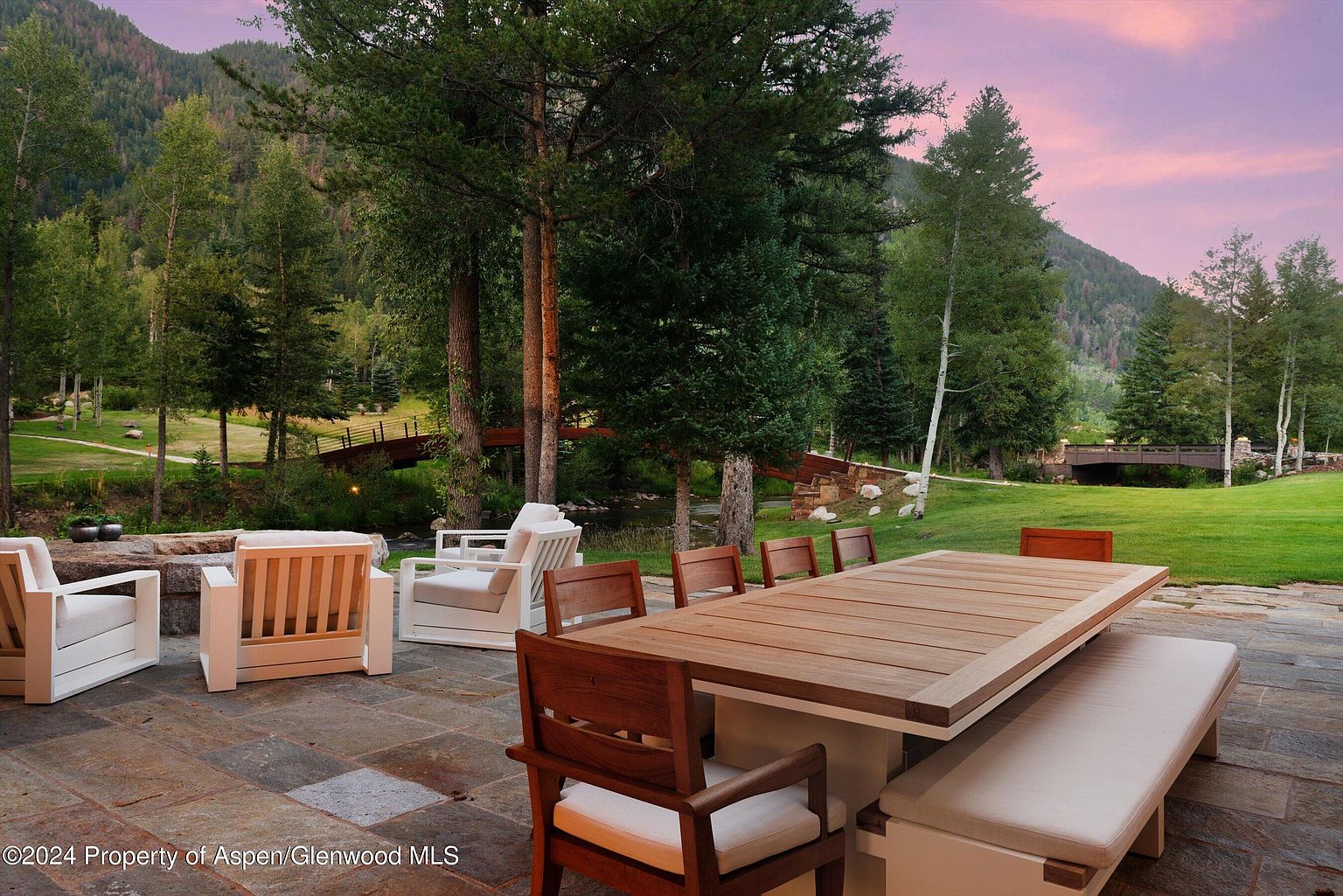 This image showcases an inviting outdoor patio area, perfect for entertaining or relaxing. The space features a large wooden dining table with bench seating and comfortable chairs, all set on a stone-tiled patio. In the background, lush greenery, mature trees, and a charming bridge create a serene and picturesque setting, enhancing the property's appeal.