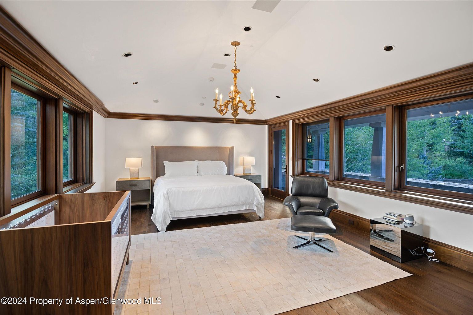 This is a primary bedroom featuring a king-size bed with a neutral-toned headboard, flanked by matching nightstands and lamps. The room is well-lit with natural light streaming through large windows framed with dark wood trim, complementing the hardwood flooring. A decorative chandelier hangs from the ceiling, adding a touch of elegance to the space, while a modern chair and ottoman offer a comfortable seating area.