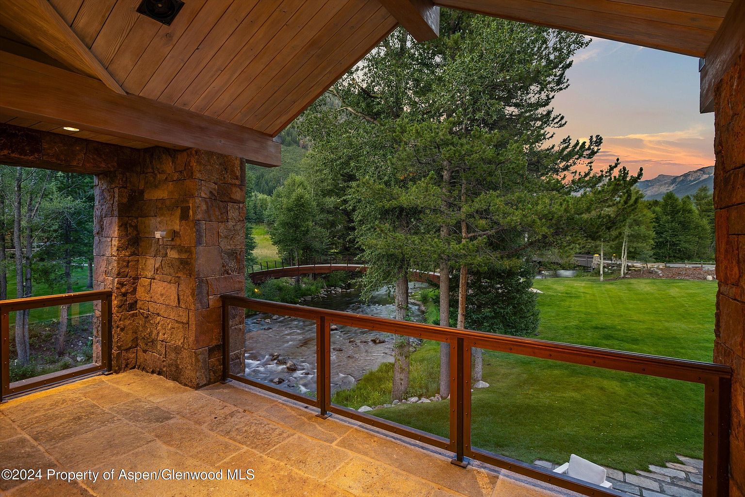 This image showcases a luxurious patio or balcony with stone flooring and a wooden railing with glass panels, offering a stunning view of a river, lush greenery, and a bridge. The structure features stone pillars and a wooden ceiling, adding to the rustic yet elegant aesthetic. The scene is bathed in soft, natural light, creating a serene and inviting atmosphere.