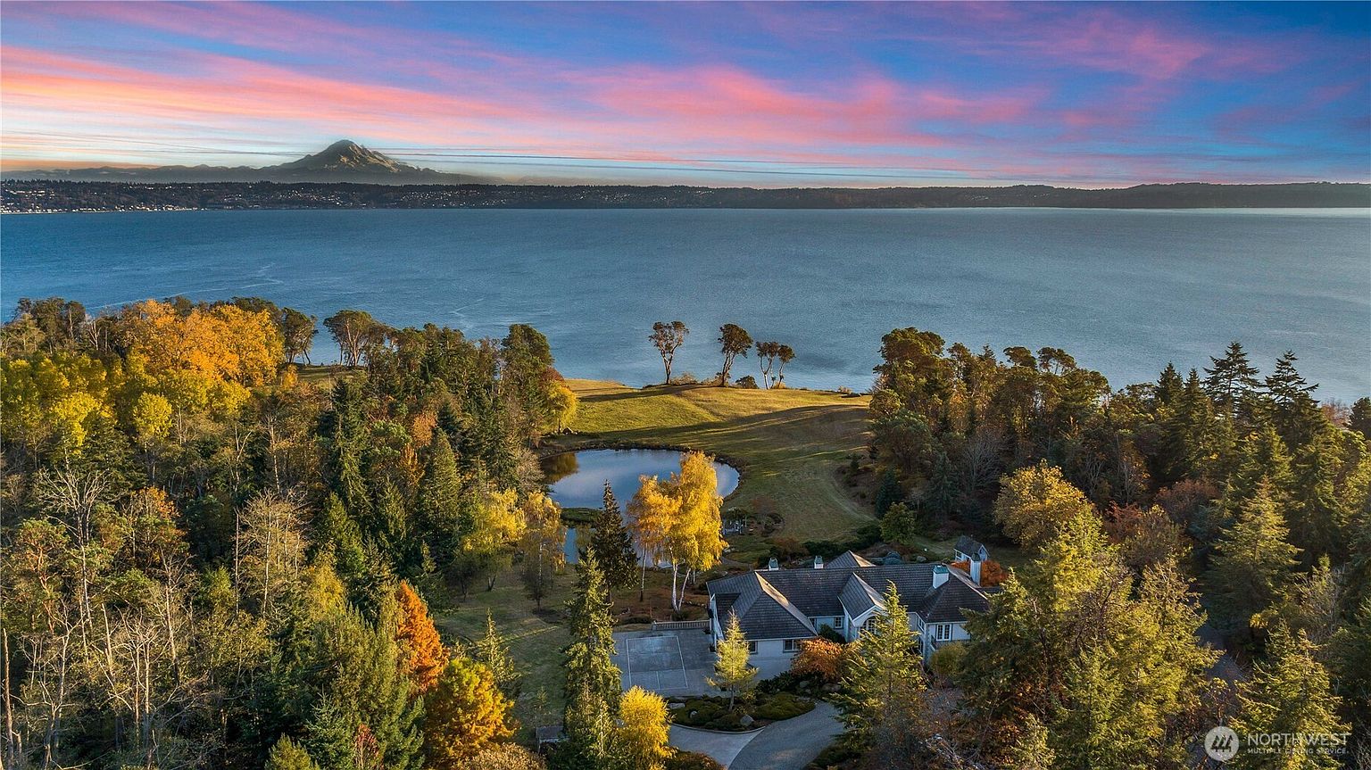 This stunning aerial perspective captures a sprawling waterfront estate nestled amidst a lush, wooded landscape during a vibrant sunset. The property features a private pond, expansive green lawns, and a multi-gabled residence, all set against the backdrop of a vast body of water and a distant, snow-capped mountain peak. The composition highlights the serene, secluded nature of the home and its exceptional integration with the natural environment.