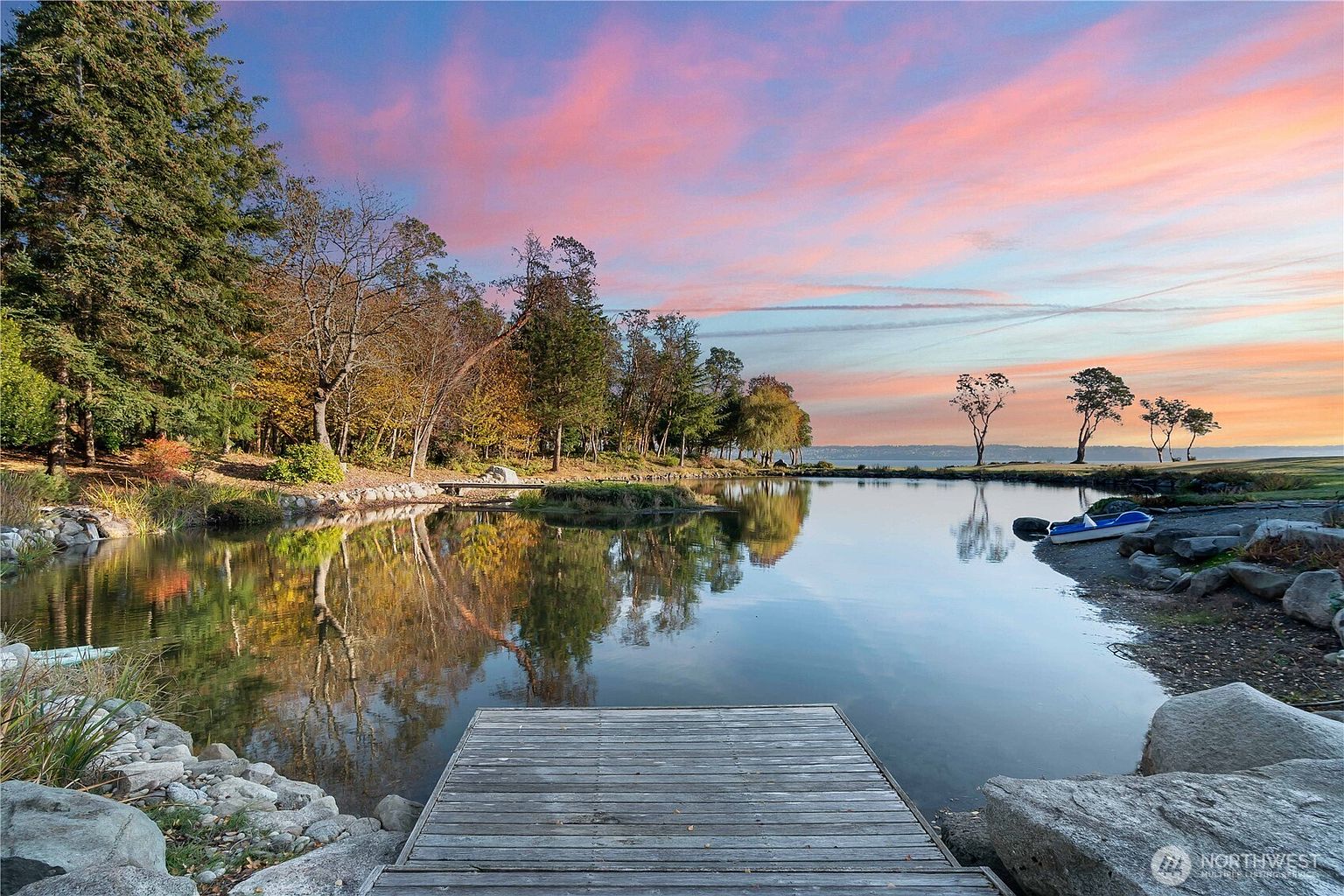 This serene waterfront property features a wooden dock extending into a calm pond, framed by lush trees and rocky landscaping. The scene is captured at sunset, with a vibrant pink and orange sky reflecting beautifully on the water's surface. A small blue boat rests on the rocky shore, enhancing the peaceful, recreational atmosphere of this expansive outdoor space.