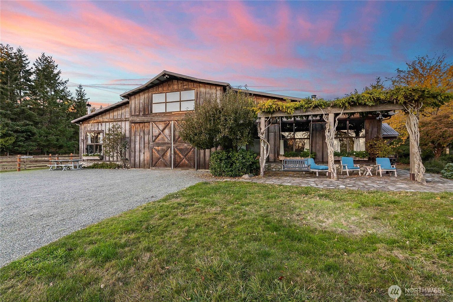 This rustic barn-style home features weathered wood siding and a prominent central sliding door, set against a vibrant sunset sky. A stone-paved patio area extends from the side, shaded by a wooden pergola draped in climbing vines and furnished with outdoor seating. The property is surrounded by a lush lawn and mature trees, creating a serene and picturesque rural atmosphere.
