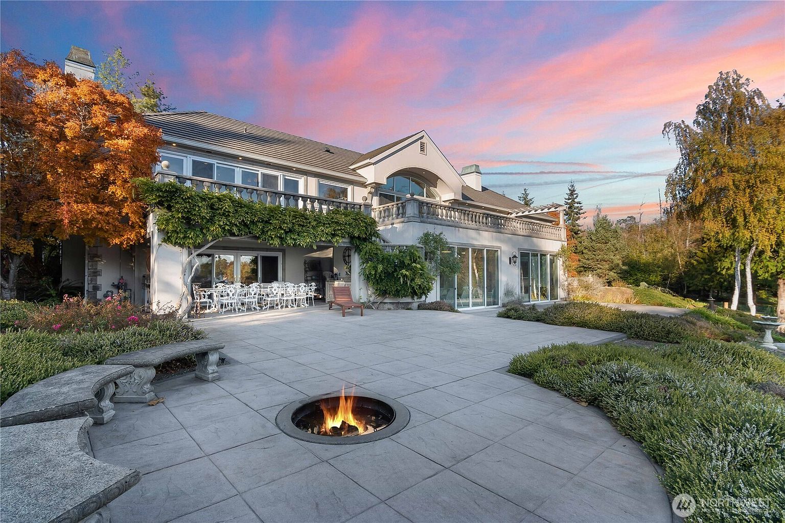 This elegant rear exterior view showcases a sprawling stone patio featuring a circular fire pit and curved stone benches, perfect for outdoor entertaining. The home features a large covered dining area draped in lush greenery, a second-story balcony with a stone balustrade, and expansive glass doors that connect the indoor and outdoor living spaces. The scene is captured at dusk, with a vibrant sunset sky providing a dramatic backdrop to the manicured landscaping and mature trees.
