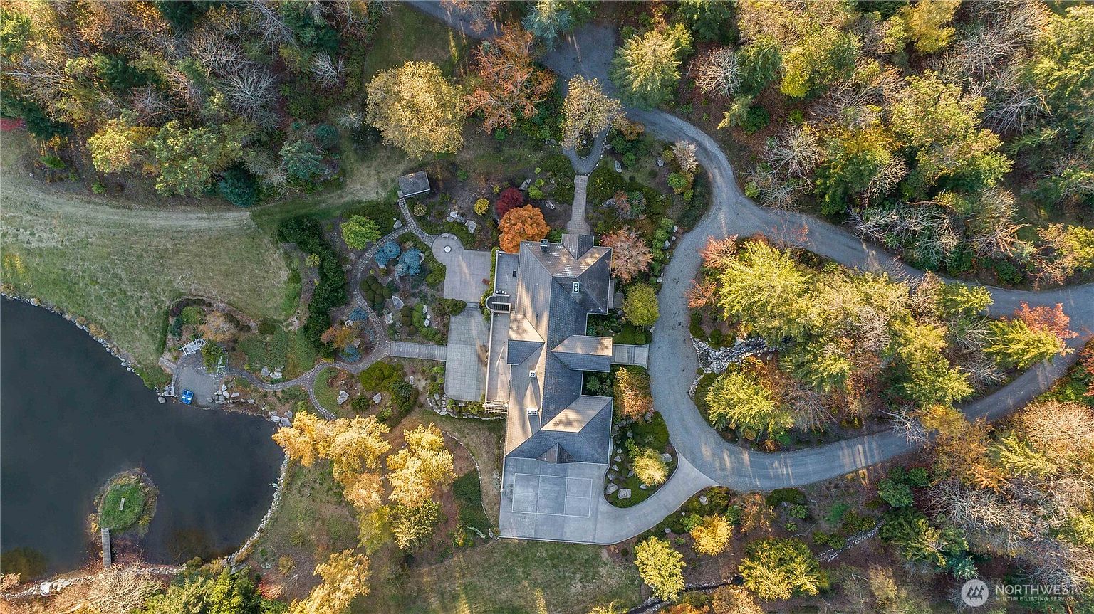 This high-angle aerial view captures a sprawling estate nestled within a lush, wooded landscape. The property features a multi-gabled residence with a dark roof, a winding gravel driveway, and an expansive, meticulously landscaped garden area that transitions into a serene pond. The perspective highlights the home's integration into the natural environment, showcasing the mature trees and peaceful outdoor living spaces.