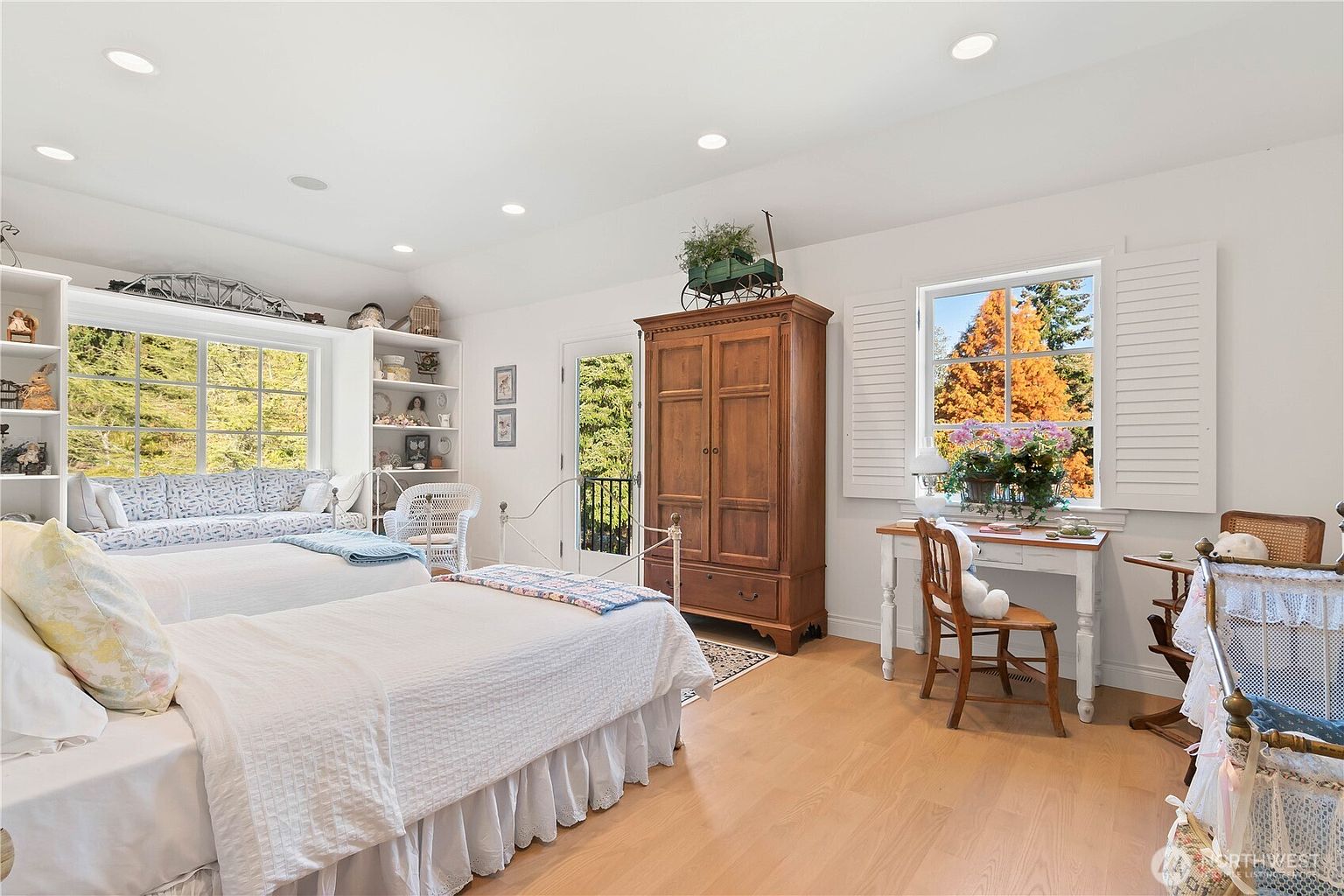 This bright and airy bedroom features two twin beds with white linens, a built-in window seat, and a classic wooden armoire. A small desk with a chair sits near a window, while a vintage-style cradle adds a charming touch to the corner. The room is finished with light wood flooring and recessed lighting, creating a warm and inviting atmosphere.
