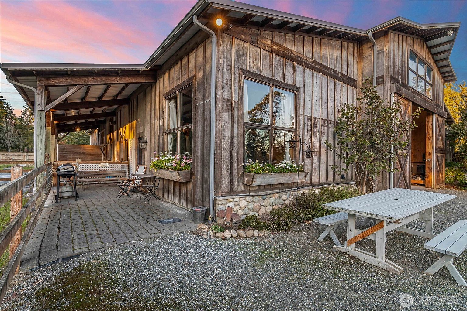 This rustic exterior view captures a charming barn-style home featuring weathered wood siding and a stone foundation. A covered patio area with stone pavers provides a cozy outdoor living space, complete with a grill, bench seating, and flower boxes beneath large windows. A picnic table sits on the adjacent gravel area, all set against a soft, sunset-lit sky that enhances the warm, inviting atmosphere of the property.