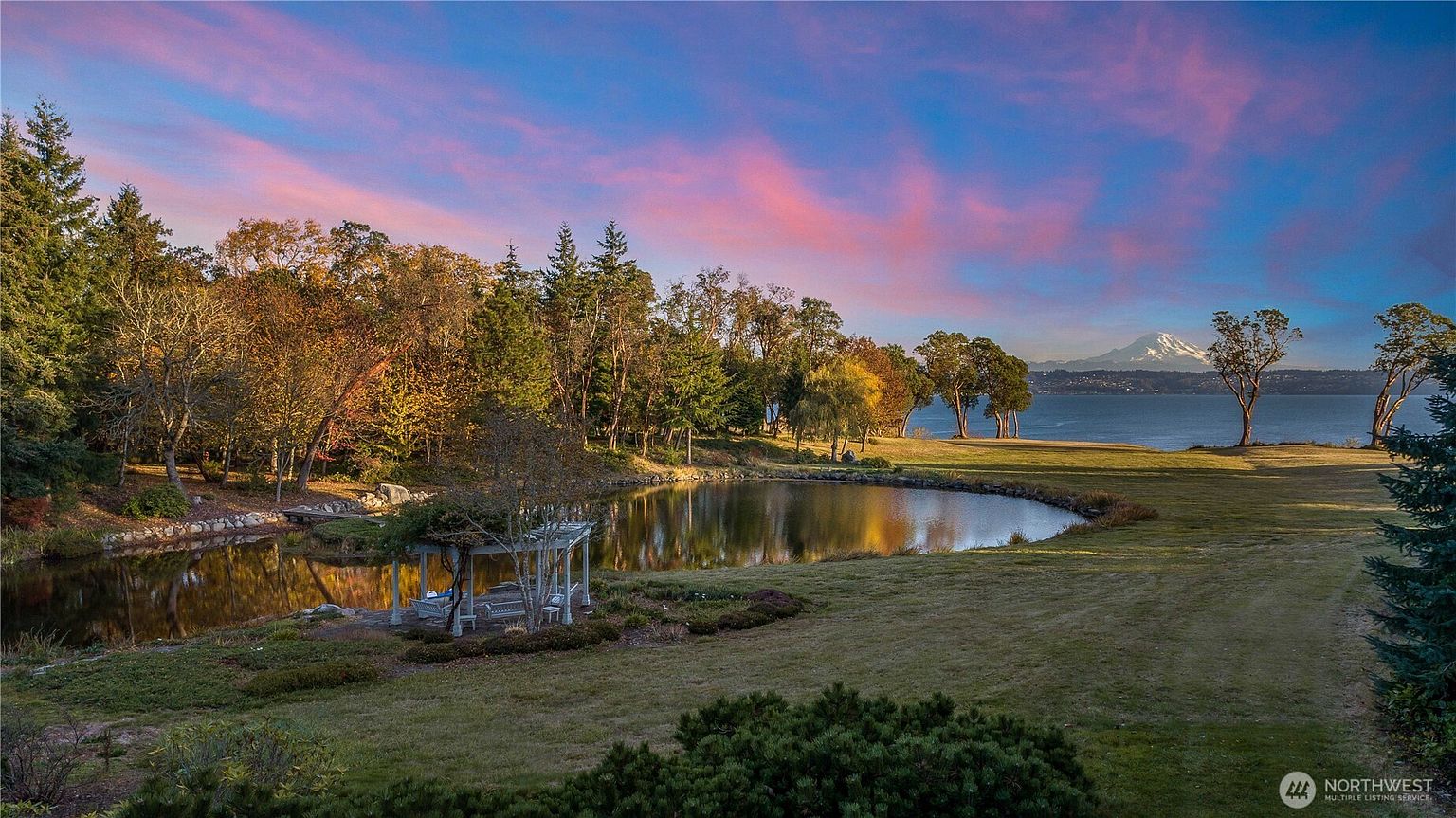 This expansive, picturesque landscape features a serene pond in the foreground, complete with a charming white pergola and a small wooden bridge. The lush green lawn stretches toward a scenic waterfront, framed by mature trees and a stunning view of a snow-capped mountain peak under a vibrant, colorful sunset sky. The perspective is captured from an elevated vantage point, emphasizing the vastness and tranquility of the property's natural setting.