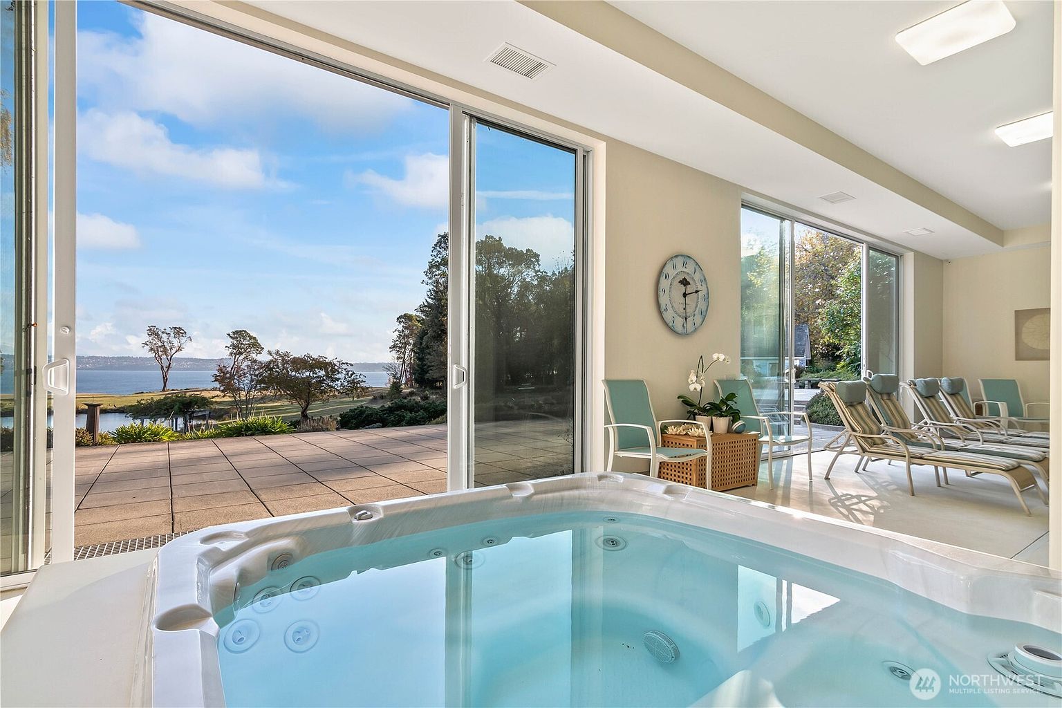 This bright, airy sunroom features a large indoor hot tub as the central focus, positioned in front of expansive sliding glass doors that offer panoramic views of the water and landscape. The space is furnished with several lounge chairs, creating a relaxing retreat that seamlessly blends indoor comfort with the outdoor scenery. The room is bathed in natural light, highlighting the clean, neutral aesthetic and the tranquil atmosphere of the space.