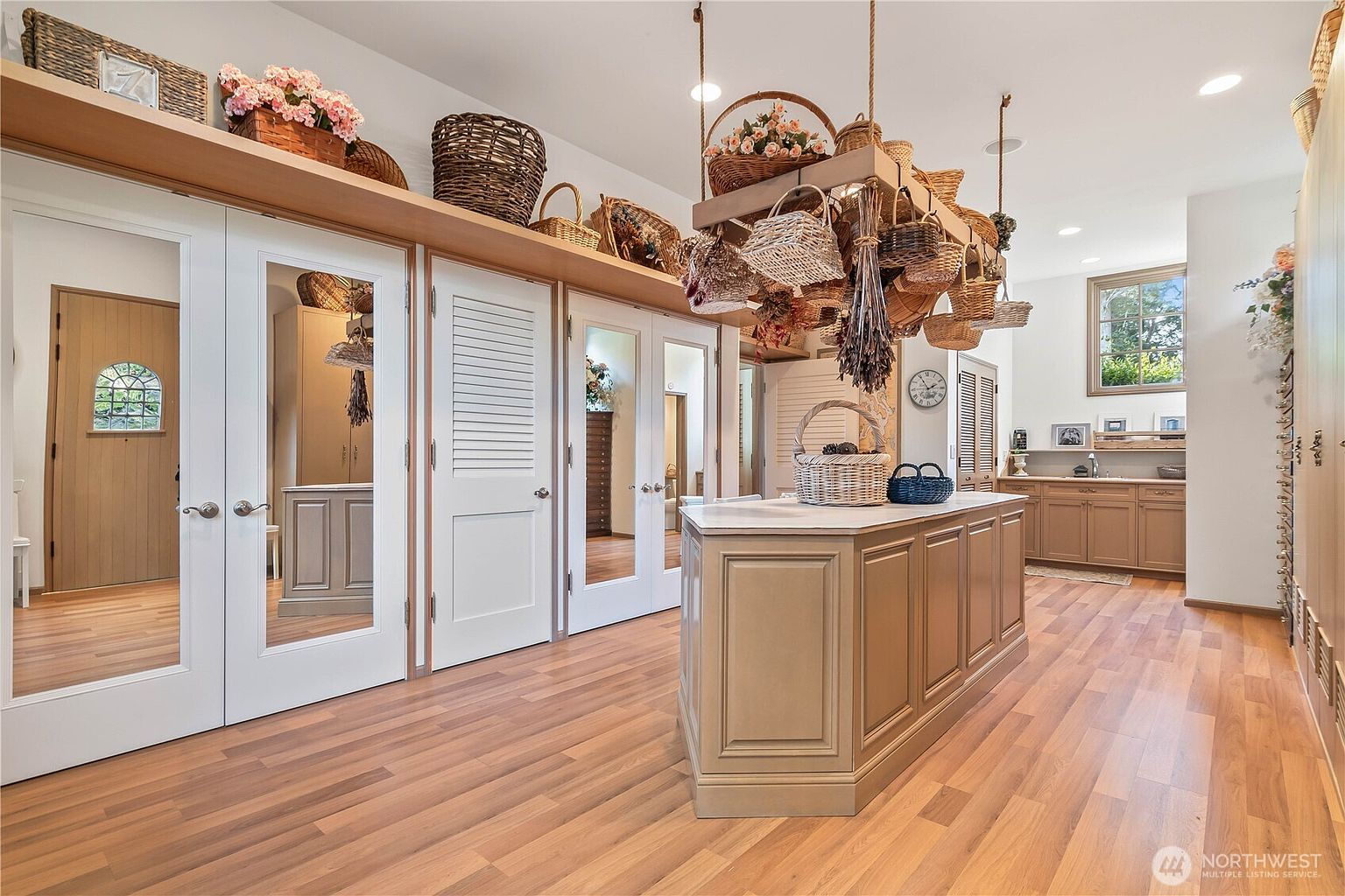 This spacious walk-in closet features a central island with a light-toned countertop and ample storage cabinetry. The room is accented by a unique hanging rack filled with various woven baskets, while mirrored closet doors reflect the surrounding space and add depth. The warm wood flooring and neutral wall tones create a bright, organized, and inviting atmosphere.