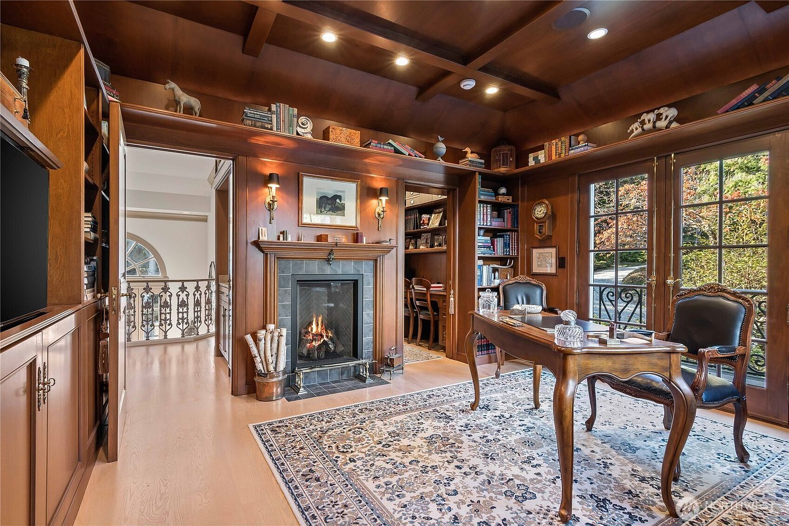 This sophisticated home office features rich wood-paneled walls, a coffered ceiling, and a traditional desk set on a large, ornate area rug. A central fireplace with a tile surround serves as a focal point, flanked by built-in bookshelves and a doorway leading to an adjacent workspace. The room is bathed in warm light from recessed fixtures and wall sconces, creating an atmosphere of classic elegance and professional comfort.