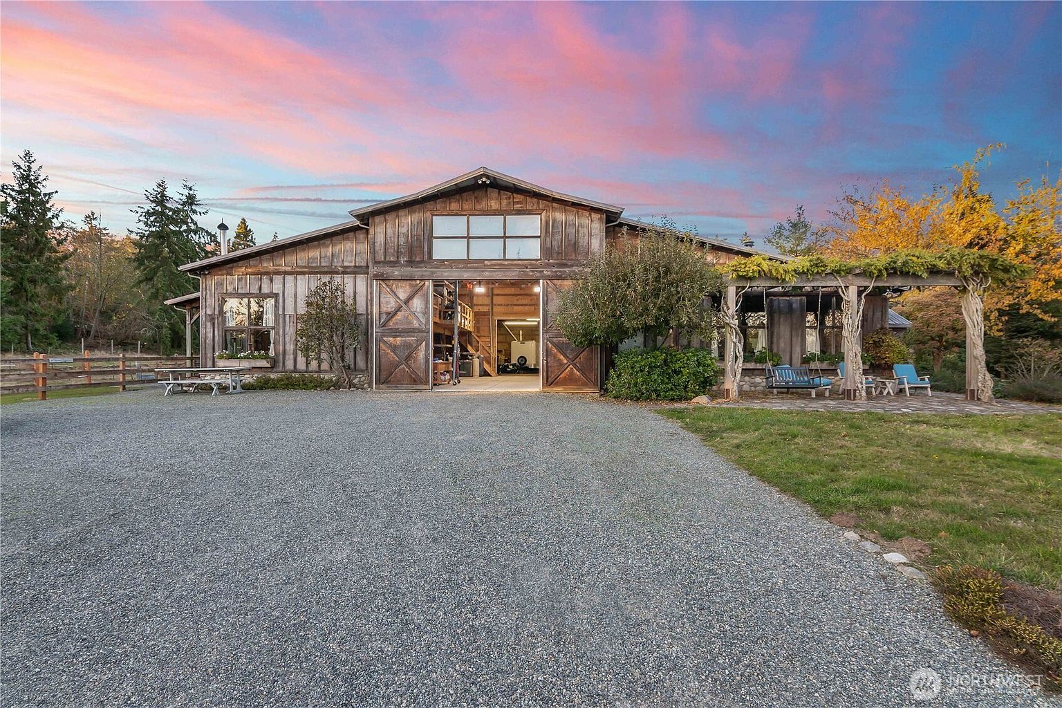 This rustic, barn-style home features weathered wood siding and a large, open central bay with sliding barn doors. A gravel driveway leads up to the structure, which is complemented by a stone-paved patio area shaded by a wooden pergola with climbing vines. The scene is captured at dusk, with a vibrant, colorful sunset sky providing a dramatic backdrop to the property's pastoral setting.