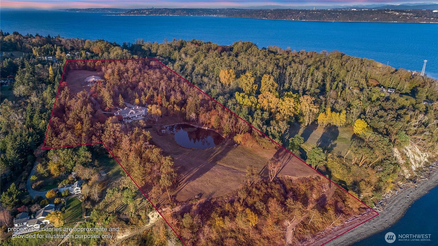 This high-angle aerial view captures a sprawling, wooded waterfront estate defined by a red boundary line. The property features a large private pond, a main residence, and a secondary structure, all nestled within a dense forest that slopes down toward a rocky beach and the expansive blue water beyond. The perspective provides a comprehensive look at the vast acreage and the unique coastal landscape of the parcel.