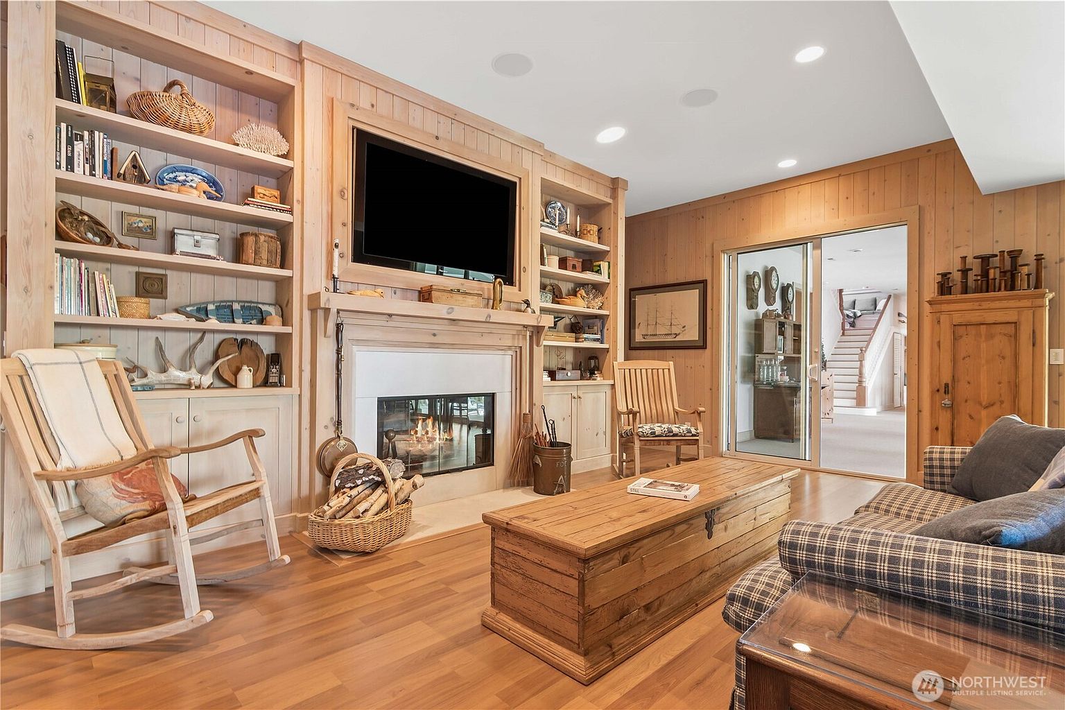 This cozy living room features extensive light wood built-in shelving flanking a central fireplace with a mounted television above the mantel. A rustic wooden trunk serves as a coffee table in the center of the room, complemented by a plaid sofa and a wooden rocking chair. The space is warm and inviting, with a sliding glass door providing a view into an adjacent hallway and staircase.