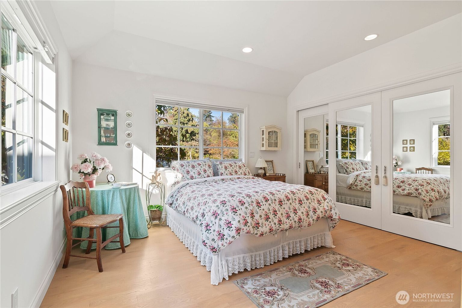 This bright and airy bedroom features light wood flooring and a vaulted ceiling, creating a spacious and welcoming atmosphere. The room is furnished with a bed dressed in floral bedding, a small round table with a teal cloth, and a wooden chair, all complemented by large windows that allow for ample natural light. A large mirrored closet door reflects the room, adding depth and enhancing the sense of space.