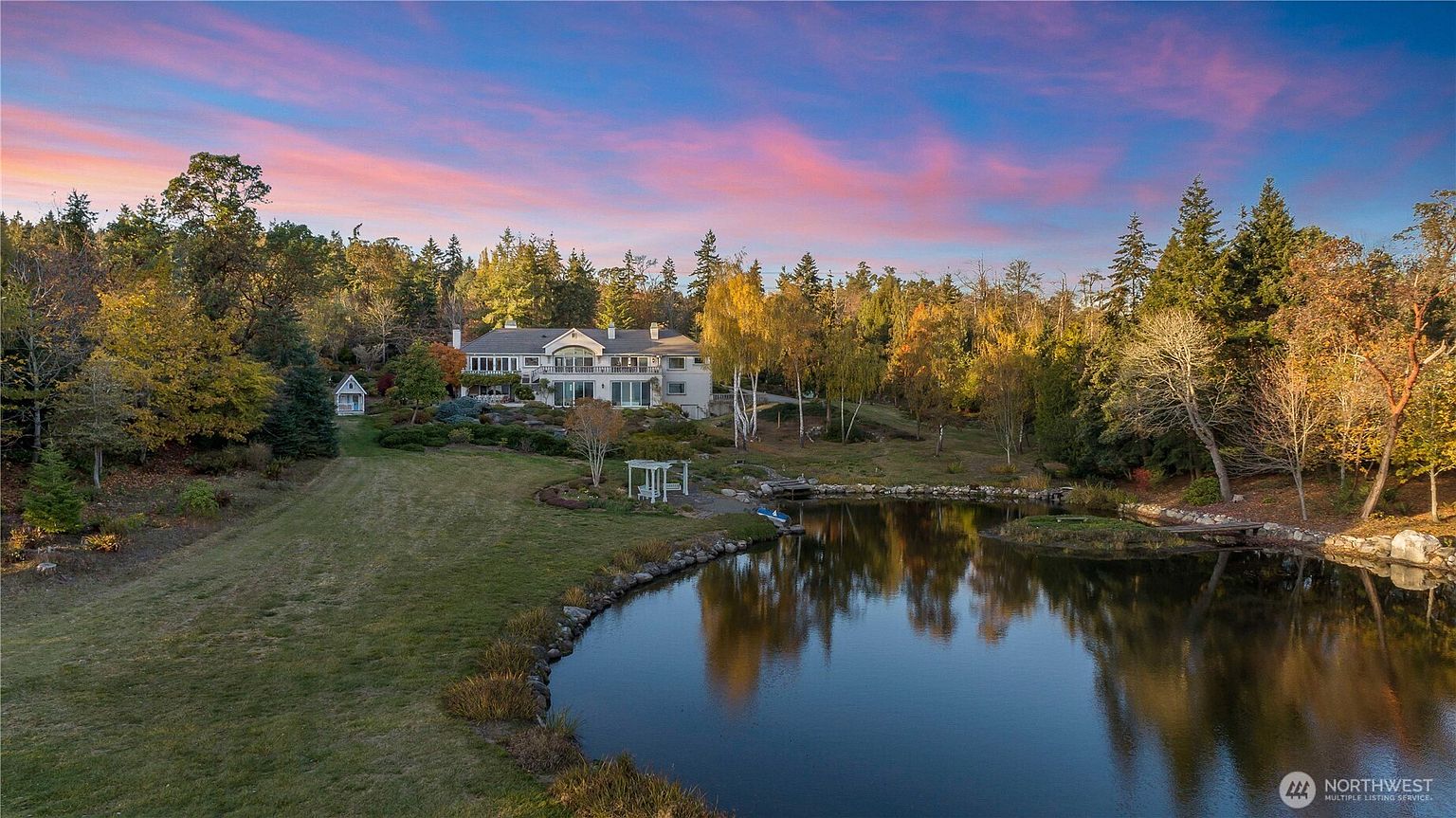 This elevated perspective captures a sprawling estate home nestled within a lush, wooded landscape at sunset. The property features a large, tranquil pond in the foreground, a manicured lawn, and a multi-level residence with a prominent balcony overlooking the grounds. The warm, vibrant colors of the sky reflect beautifully in the water, creating a serene and picturesque atmosphere.