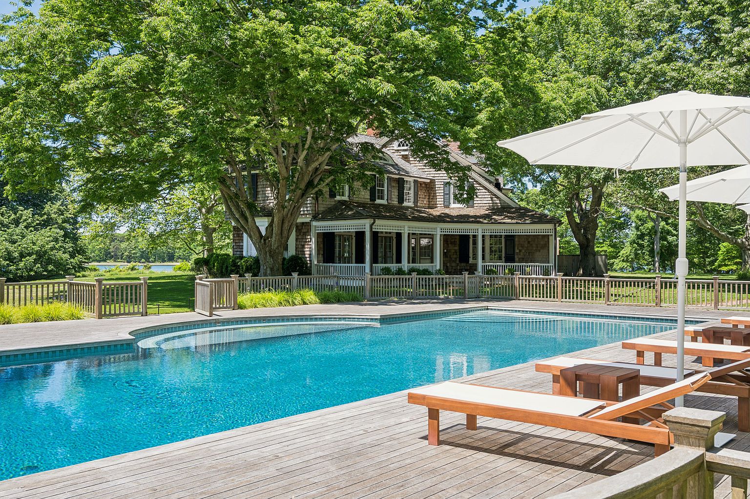 This serene outdoor space features a large, custom-shaped swimming pool set into a spacious wooden deck. The scene is framed by a classic shingle-style house with a covered porch, surrounded by lush, mature trees and a manicured lawn. Several wooden lounge chairs with white cushions are arranged under a large white umbrella, creating an inviting atmosphere for relaxation and leisure.