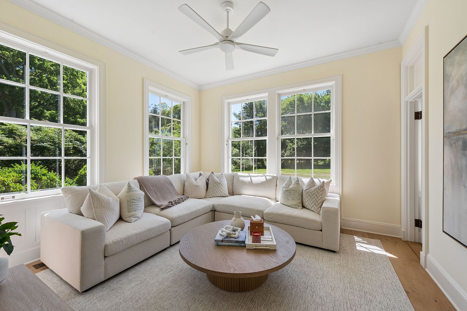 This bright and airy living room features a large, neutral-toned sectional sofa arranged in an L-shape, centered around a round, light wood coffee table. The space is illuminated by multiple large windows that provide ample natural light and views of the surrounding greenery. A white ceiling fan hangs above, and the room is finished with light-colored walls and wood flooring, creating a clean, inviting, and contemporary atmosphere.