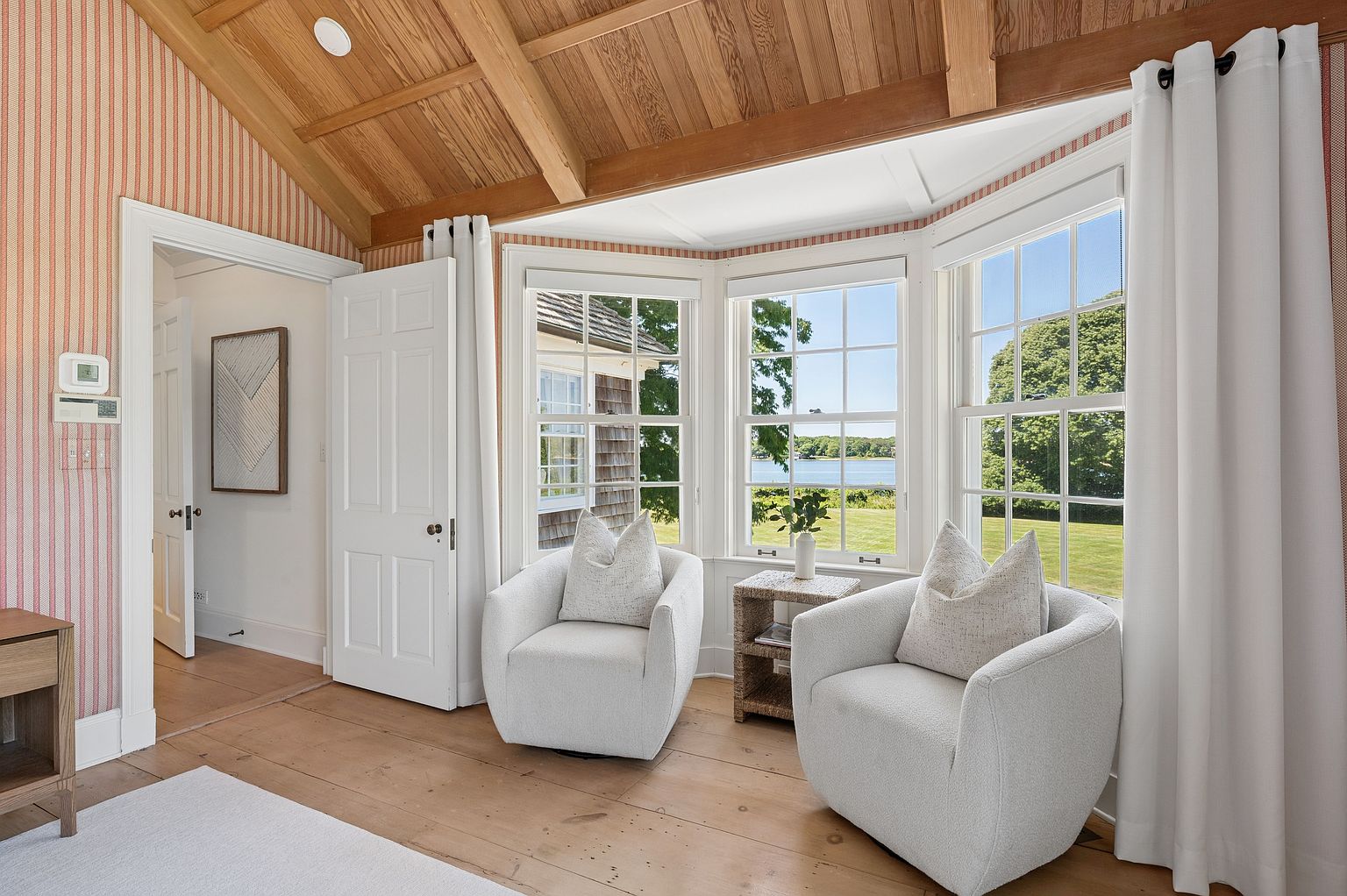 This bright sunroom features a vaulted wood-paneled ceiling with exposed beams and charming striped wallpaper. A bay window arrangement provides expansive views of the lush green landscape and water beyond, while two plush, light-colored armchairs create a cozy seating area. The space is finished with light wood flooring and a small woven side table, offering a serene and inviting atmosphere.