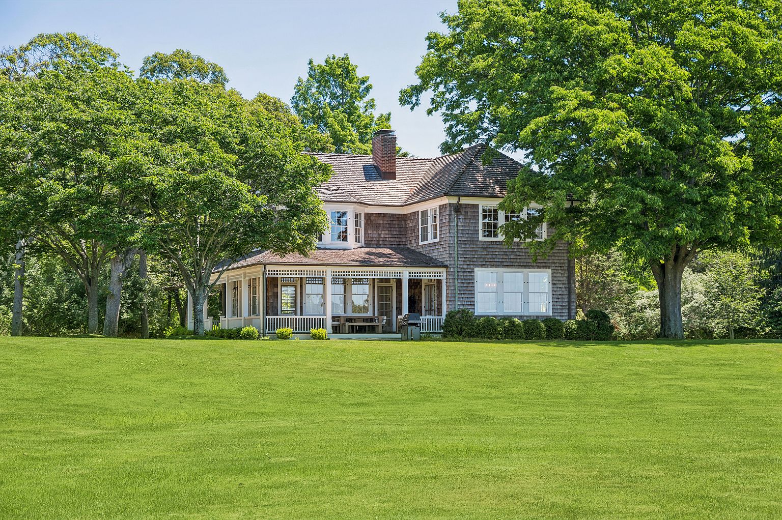 This classic shingle-style home is set against a backdrop of lush, mature trees and a sprawling, manicured lawn. The rear facade features a covered porch with white columns, multiple windows, and a prominent brick chimney rising from the roofline. The overall impression is one of timeless elegance and peaceful, private living in a well-maintained landscape.