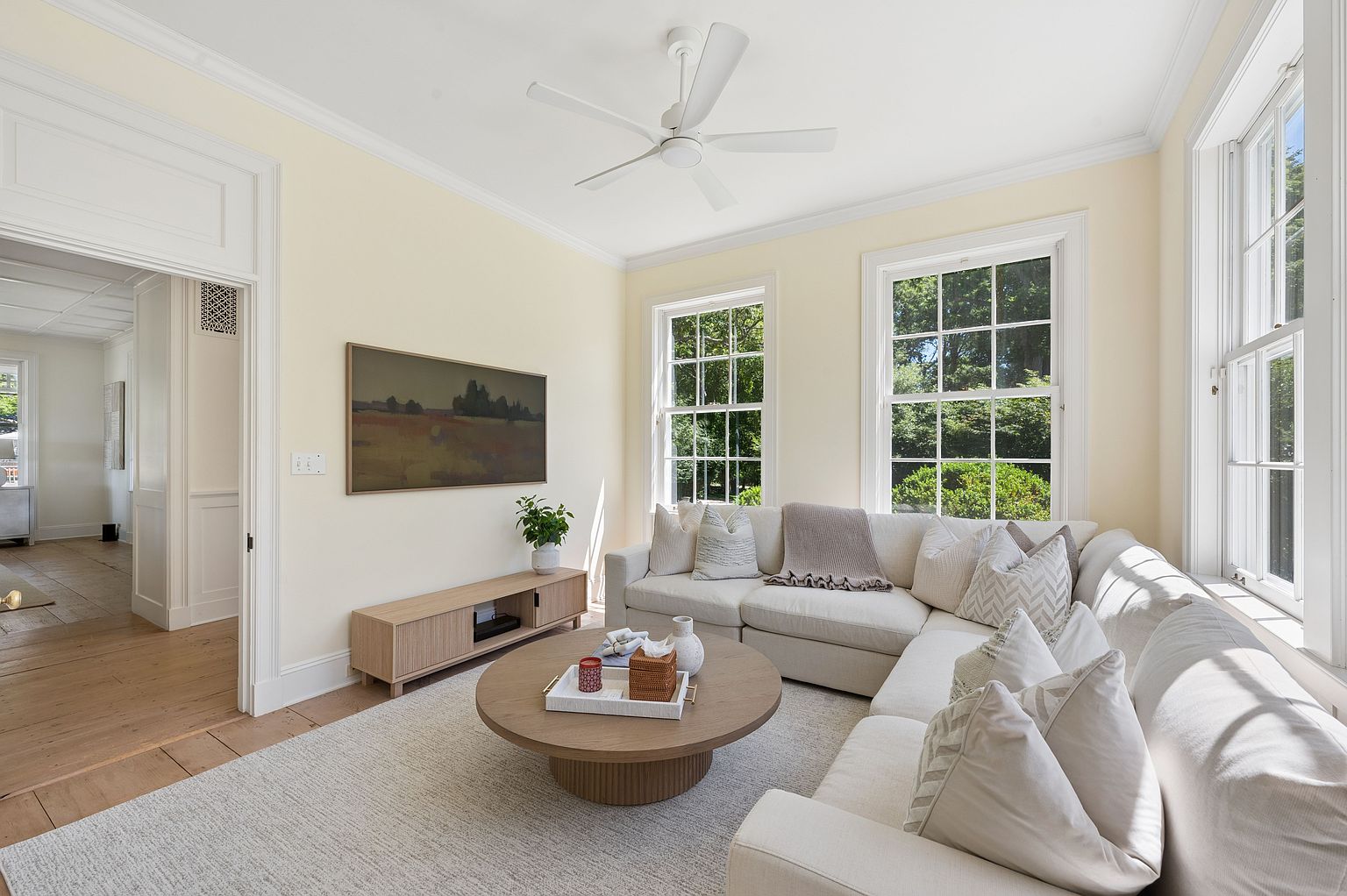 This bright and airy living room features a large, light-colored sectional sofa arranged around a circular wooden coffee table on a textured area rug. The space is accented by a light wood media console, a piece of landscape art on the wall, and a white ceiling fan, all set against soft cream-colored walls. Large, multi-pane windows allow ample natural light to fill the room, creating a welcoming and comfortable atmosphere.