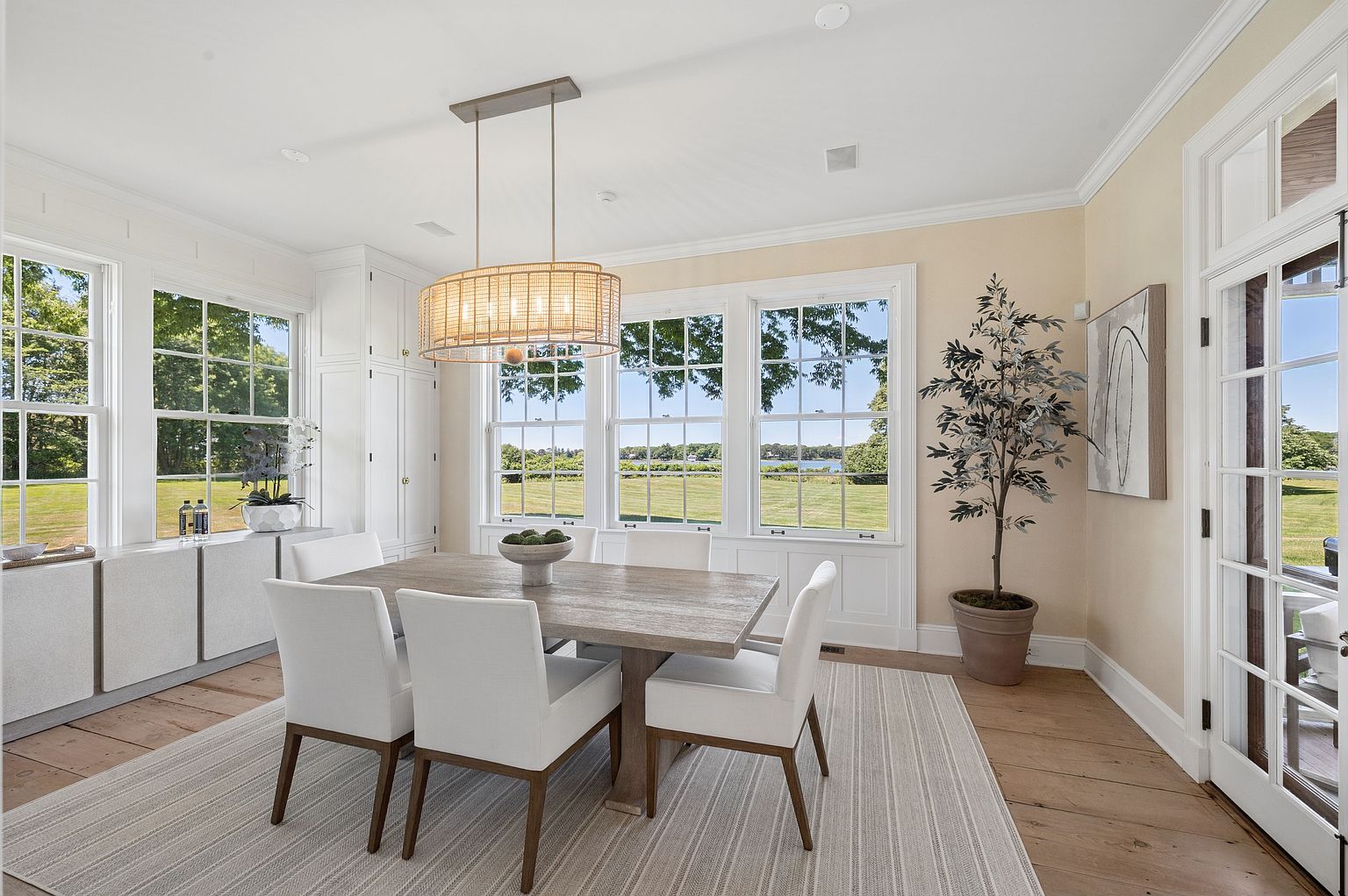 This bright and airy dining room features a light wood table surrounded by four white upholstered chairs, set atop a neutral striped area rug. A woven drum pendant light hangs above the table, while large windows provide expansive views of the lush green lawn and water beyond. The space is accented by a potted tree in the corner and built-in cabinetry, creating a clean, coastal-inspired aesthetic.