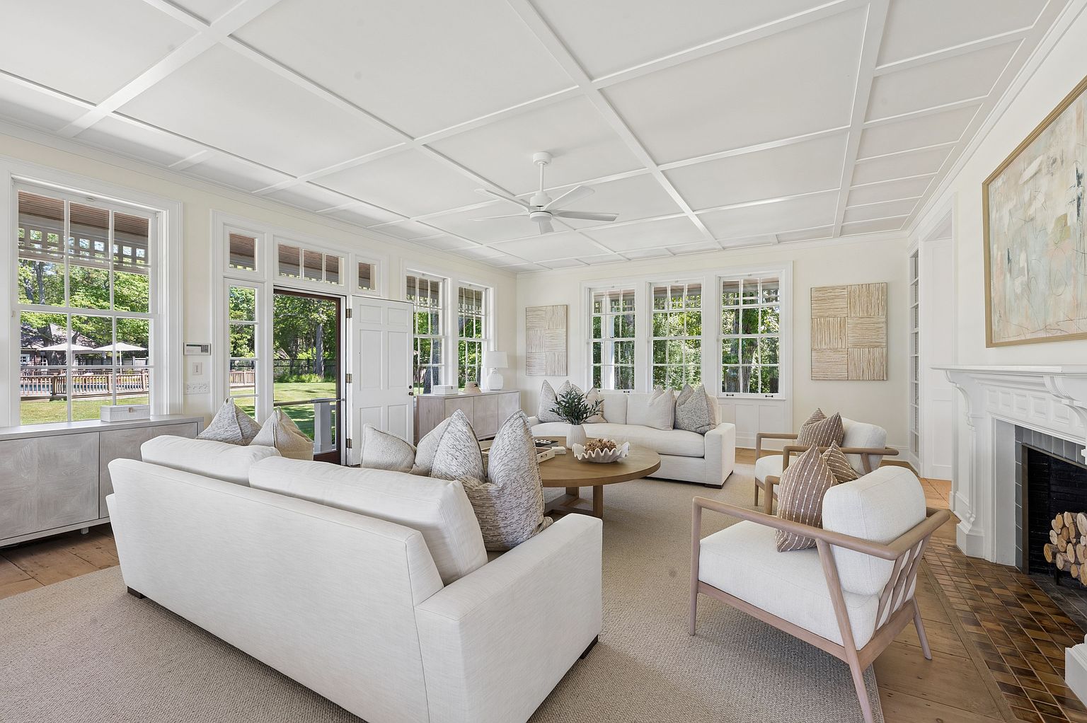 This bright and airy living room features a coffered ceiling and a neutral color palette, creating a sophisticated and inviting atmosphere. The space is furnished with two plush sofas and two accent chairs arranged around a round wooden coffee table, all resting on a large textured area rug. A classic fireplace with a white mantel serves as a focal point on the right, while large windows provide abundant natural light and views of the lush green exterior.