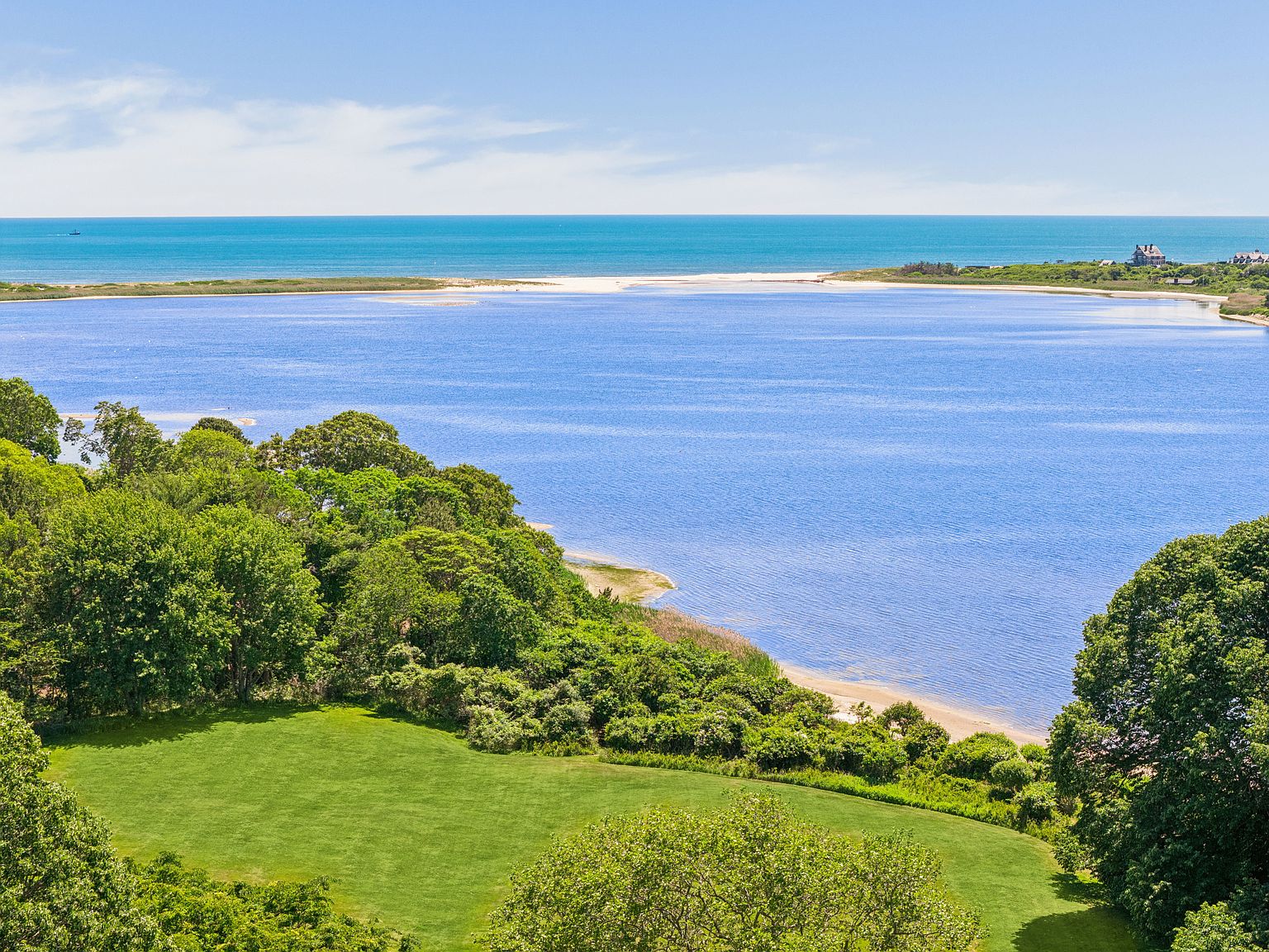 This elevated aerial perspective captures a serene coastal landscape featuring a lush green lawn that transitions into a dense tree line bordering a calm, blue tidal inlet. Beyond the inlet, a sandy spit separates the water from the vast, open ocean under a clear, bright sky. The scene conveys a sense of tranquility and privacy, highlighting the property's prime waterfront location and natural surroundings.
