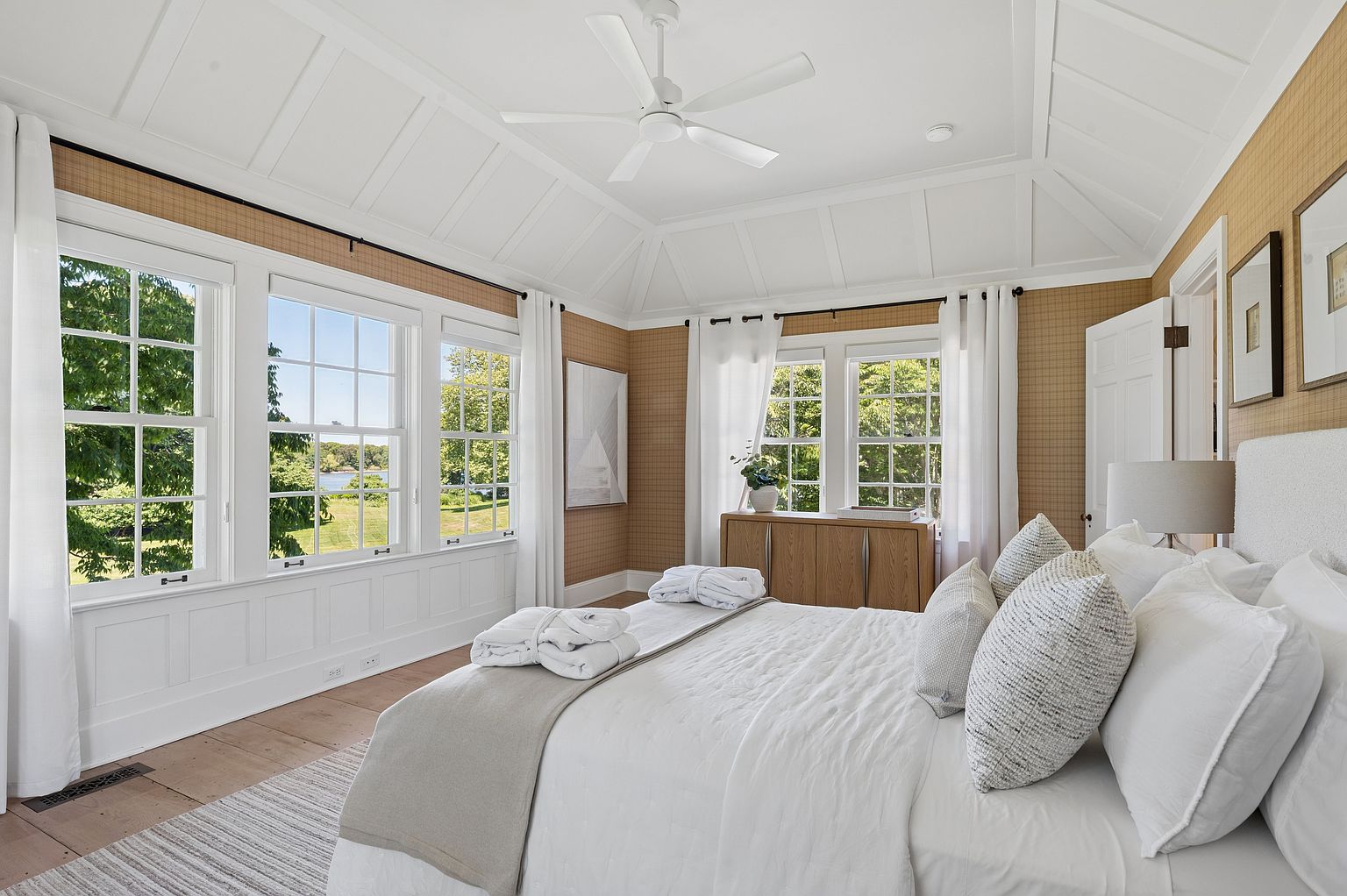This bright and airy primary bedroom features a vaulted ceiling with white paneling and a central ceiling fan. The room is decorated with warm-toned textured wallpaper, large windows that offer a scenic view of the outdoors, and a plush bed dressed in white linens. A light wood cabinet sits beneath the windows, adding a touch of natural warmth to the sophisticated, neutral-toned space.