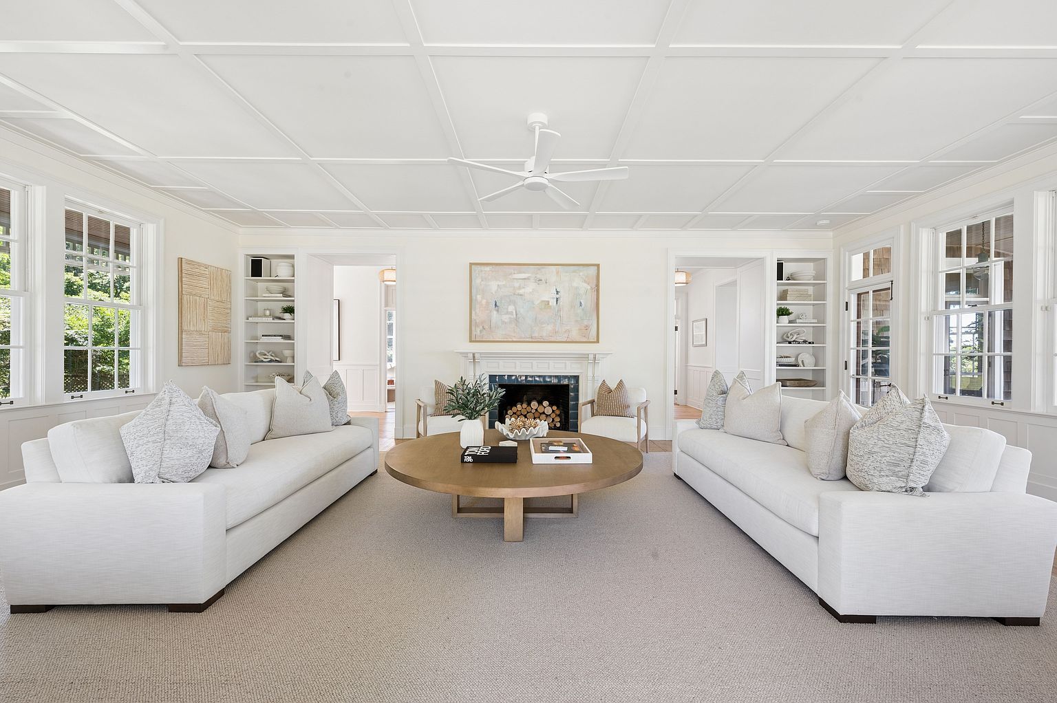 This bright and symmetrical living room features a coffered ceiling and a central fireplace with a dark tile surround and a white mantel. Two large, light-colored sofas face each other across a round wooden coffee table, creating a balanced and inviting seating area. Built-in shelving units flank the doorways on either side of the fireplace, while large windows on both walls allow for ample natural light.