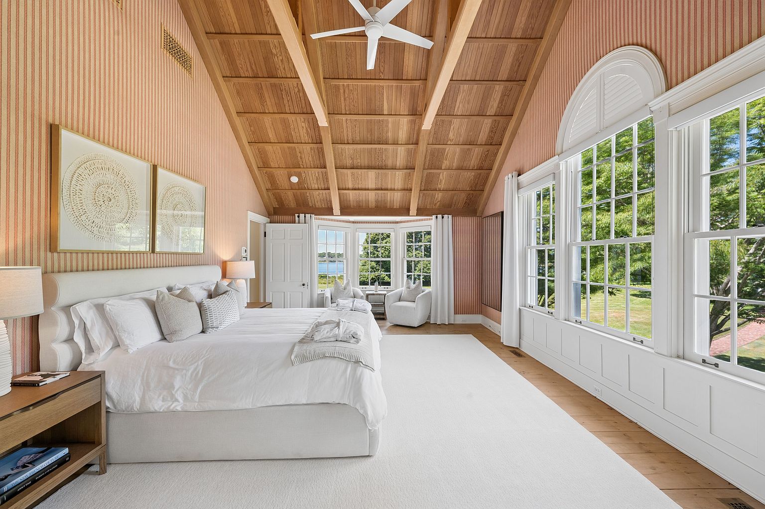 This spacious primary bedroom features a vaulted wood-paneled ceiling with exposed beams and a central ceiling fan. The room is decorated with soft pink striped wallpaper, a large white bed, and a plush white area rug, creating a serene and elegant atmosphere. Large windows with white trim provide ample natural light and views of the surrounding greenery, while a cozy seating area is tucked into the bay window alcove.