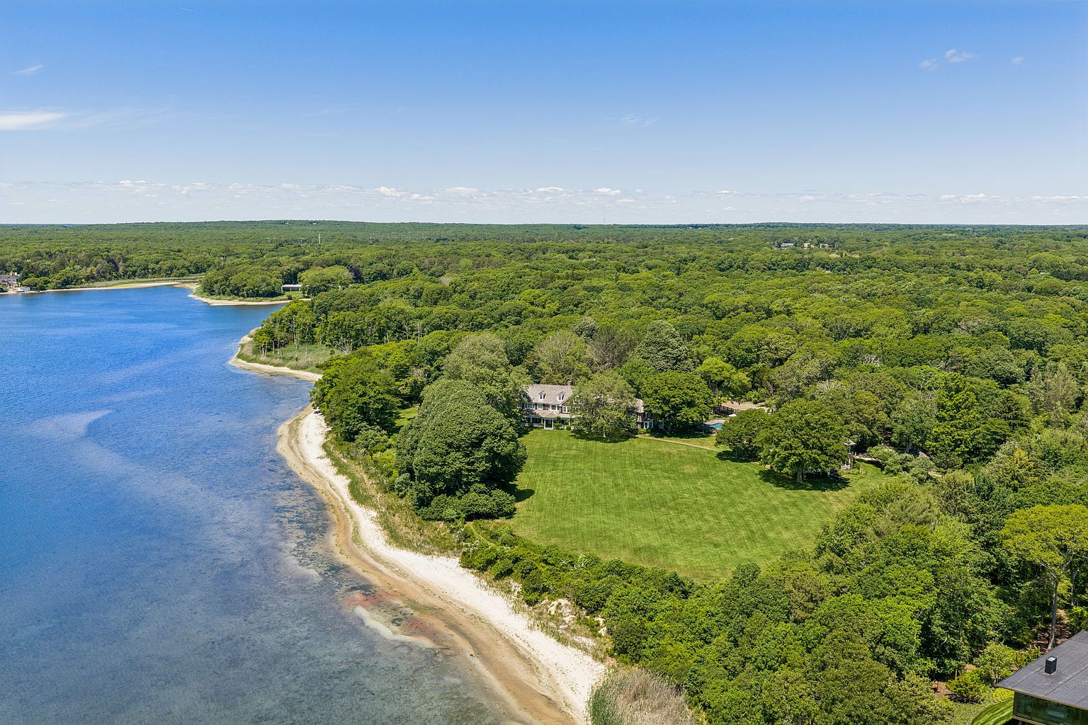 This high-angle aerial perspective captures a sprawling waterfront estate nestled within a dense, lush forest. The property features a large, manicured green lawn that transitions into a sandy shoreline along a calm, expansive body of water. The main residence is positioned centrally, surrounded by mature trees, offering a serene and private coastal retreat.