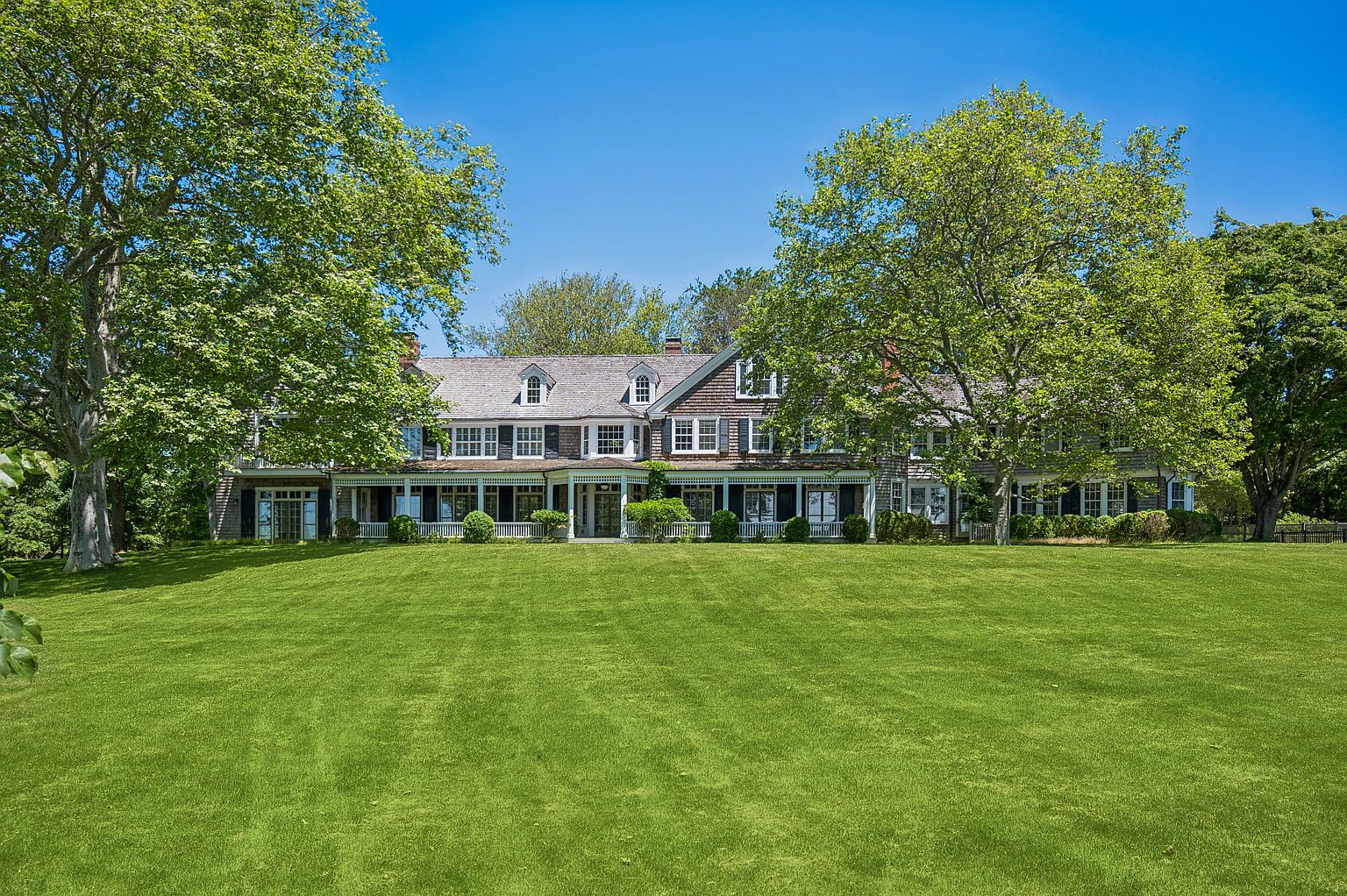 This expansive estate features a classic shingle-style residence set against a backdrop of mature, lush trees and a vast, manicured lawn. The home showcases a long, covered porch with white railings that spans the length of the rear facade, complemented by multiple dormer windows on the roofline. The perspective is a wide, eye-level shot that emphasizes the grand scale of the property and its serene, park-like setting.