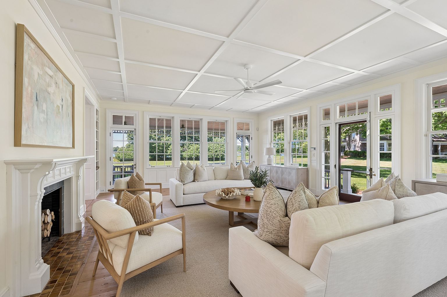 This bright and airy living room features a coffered ceiling, light-toned walls, and a large, round wooden coffee table centered on a neutral area rug. The space is furnished with two plush white sofas and two matching armchairs, all accented with textured throw pillows. A traditional fireplace with a white mantel and patterned tile hearth serves as a focal point, while large windows and glass doors provide abundant natural light and views of the surrounding greenery.