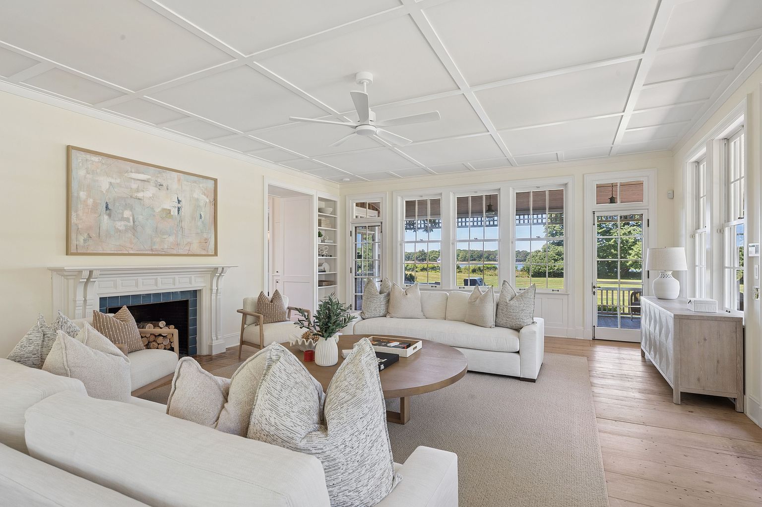 This bright and airy living room features a coffered ceiling, light wood flooring, and a neutral color palette that creates a serene atmosphere. A classic fireplace with a white mantel serves as a focal point, complemented by comfortable seating arrangements and a large round coffee table. Large windows and glass-paned doors provide abundant natural light and offer a scenic view of the surrounding landscape.