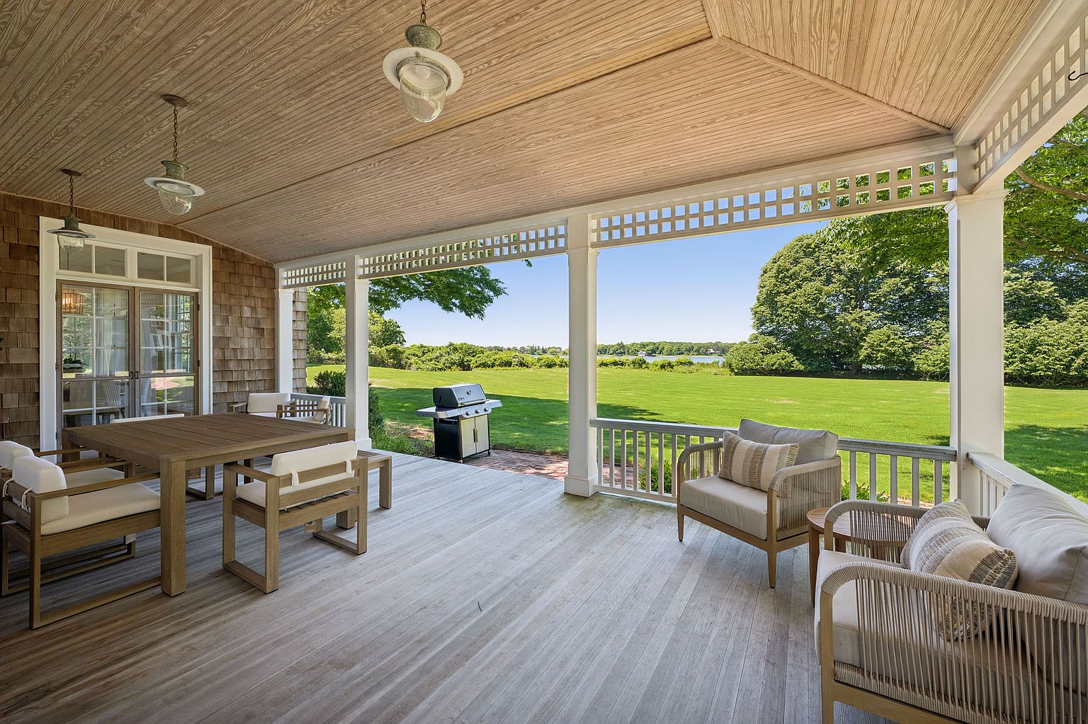 This expansive covered porch features light-toned wood decking and a classic lattice-trimmed ceiling, creating a serene outdoor living space. The area is furnished with a wooden dining set and comfortable lounge chairs, all oriented toward a sprawling, manicured lawn and distant water views. The open-air design seamlessly blends indoor comfort with the natural beauty of the surrounding landscape.