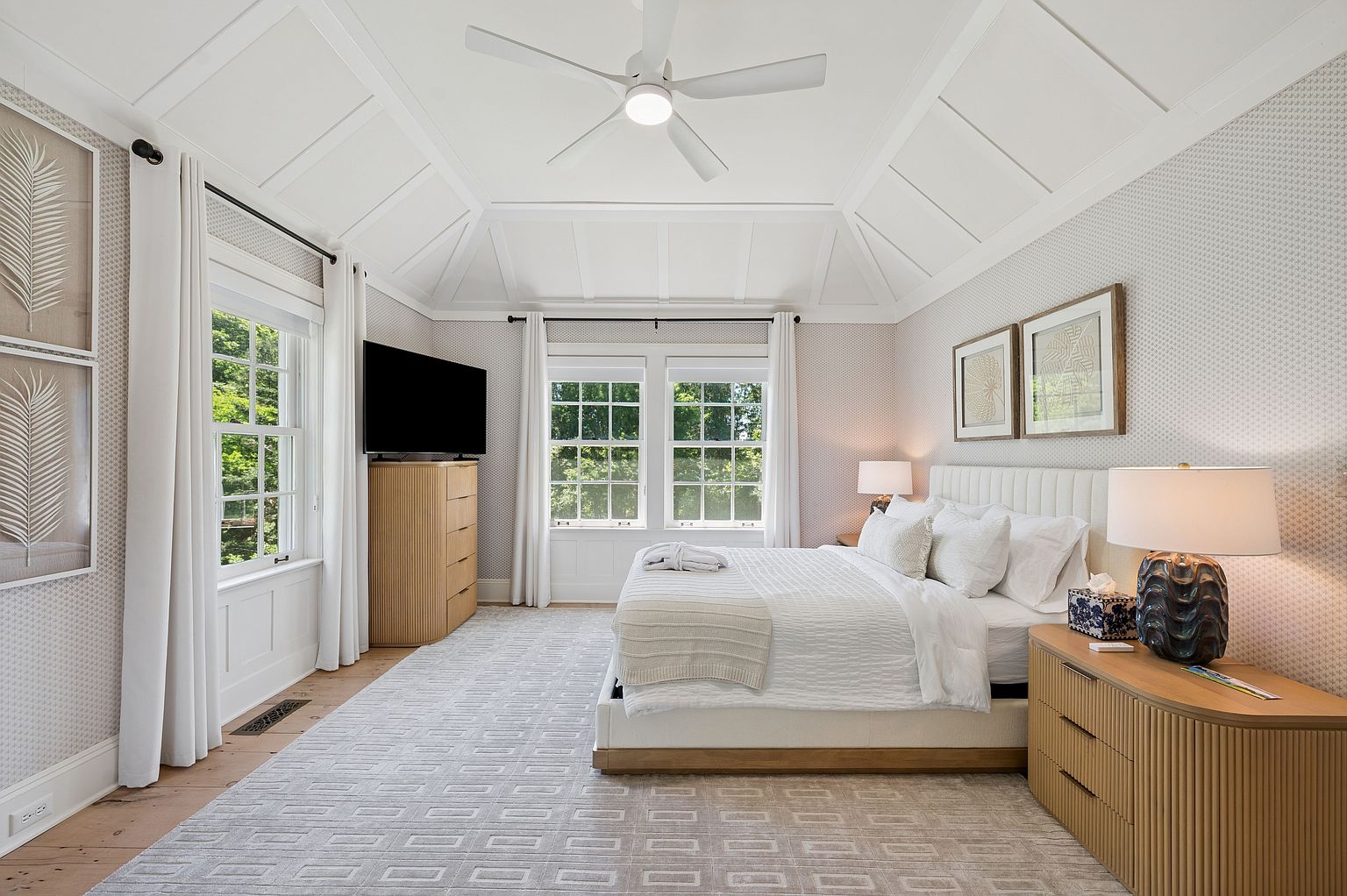 This bright and airy primary bedroom features a vaulted ceiling with white paneling and a central ceiling fan. The room is furnished with a king-sized bed, two light wood dressers with a fluted texture, and a patterned area rug that anchors the space. Large windows with white curtains allow for ample natural light, creating a serene and sophisticated atmosphere.
