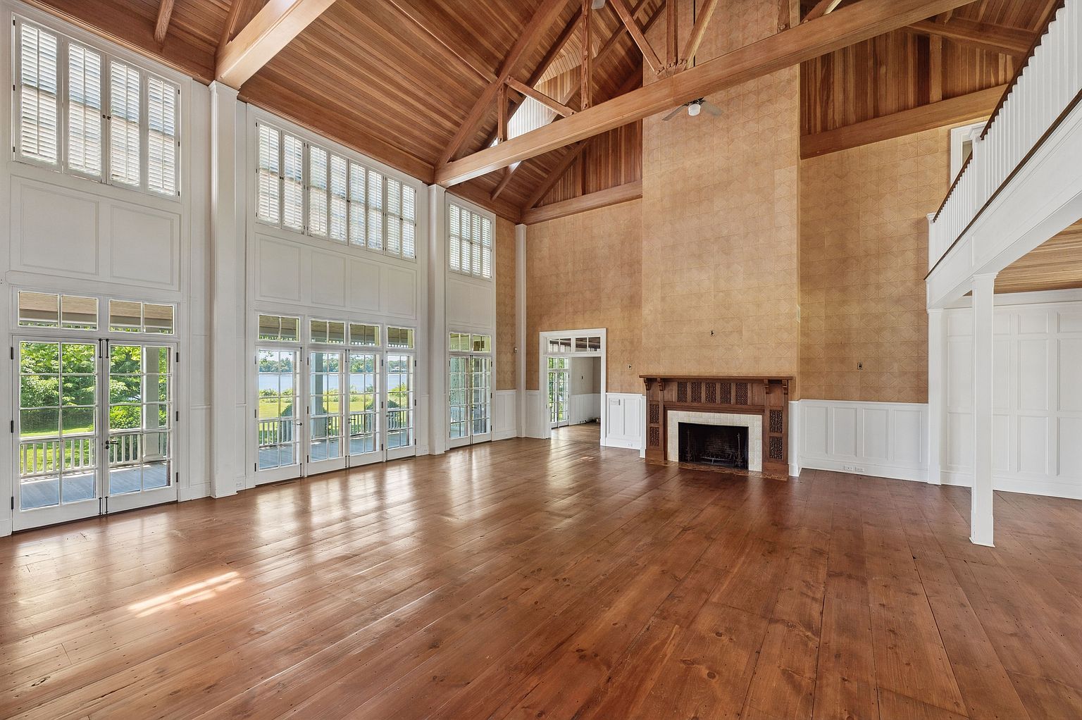 This expansive living room features a dramatic vaulted ceiling with exposed timber trusses and warm wood paneling. The space is anchored by a prominent fireplace with a detailed wood mantel and a textured, light-colored surround that extends high up the wall. Large, multi-paned windows and glass doors provide abundant natural light and views of the outdoors, while the polished wood flooring adds a sense of traditional elegance to the open, airy layout.