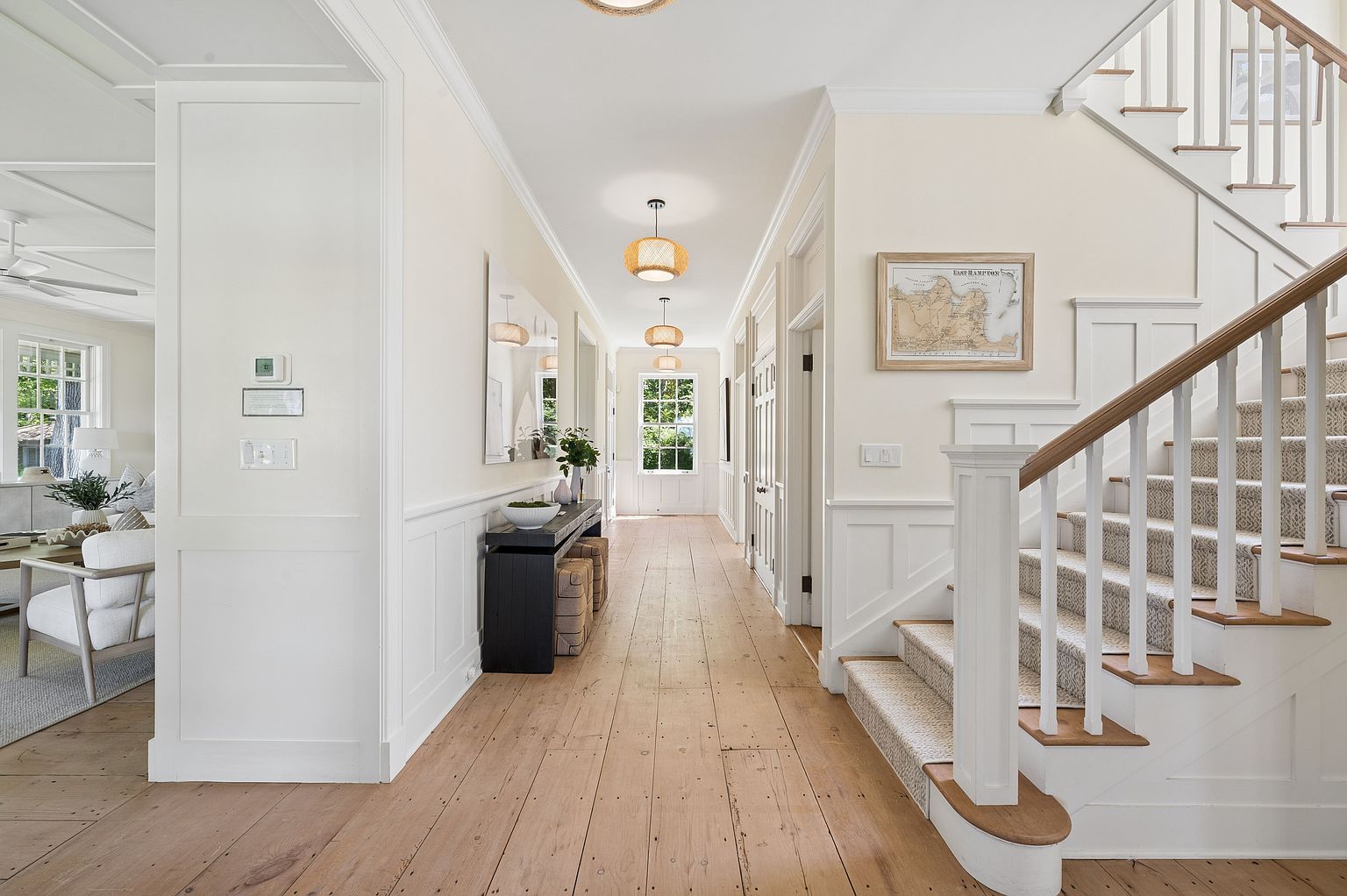 This bright and airy hallway features light wood flooring and crisp white wainscoting, creating a classic coastal aesthetic. A staircase with a patterned runner and a wooden handrail leads upward, while a long console table sits against the wall, accented by a framed map. The perspective looks down the length of the corridor toward a window at the far end, illuminated by two woven pendant lights.