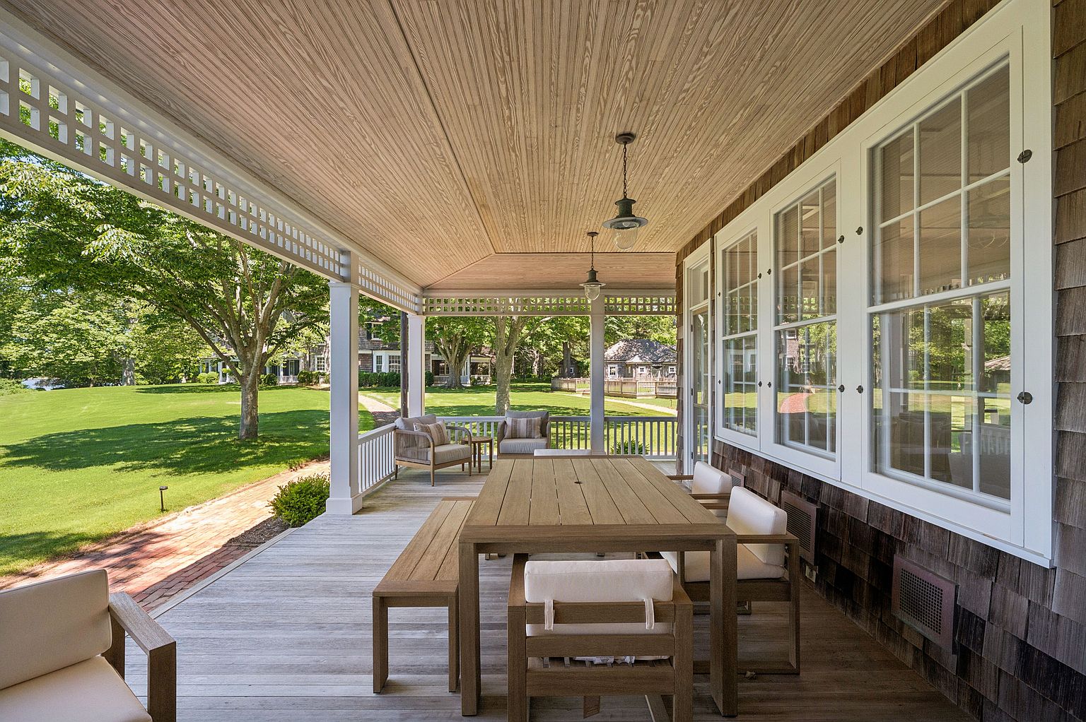 This expansive covered porch features a light wood-toned deck and a matching wood-paneled ceiling, creating a warm and inviting outdoor living space. The area is furnished with a long dining table, a bench, and chairs, all set against a backdrop of large, multi-pane windows and classic cedar shake siding. The perspective looks out over a lush, manicured green lawn and mature trees, emphasizing a serene and private residential setting.