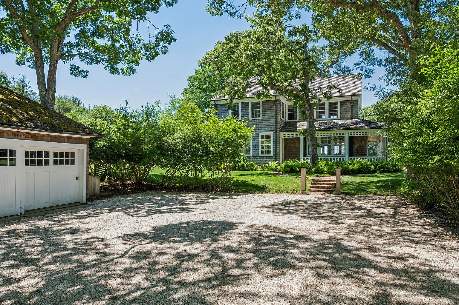 This image captures a charming, shingle-style home nestled in a lush, wooded setting, viewed from a gravel driveway. The property features a detached garage on the left and a welcoming covered porch leading to the main entrance, framed by mature trees and vibrant green landscaping. The dappled sunlight filtering through the canopy creates a serene and inviting atmosphere, highlighting the classic architectural details of the residence.