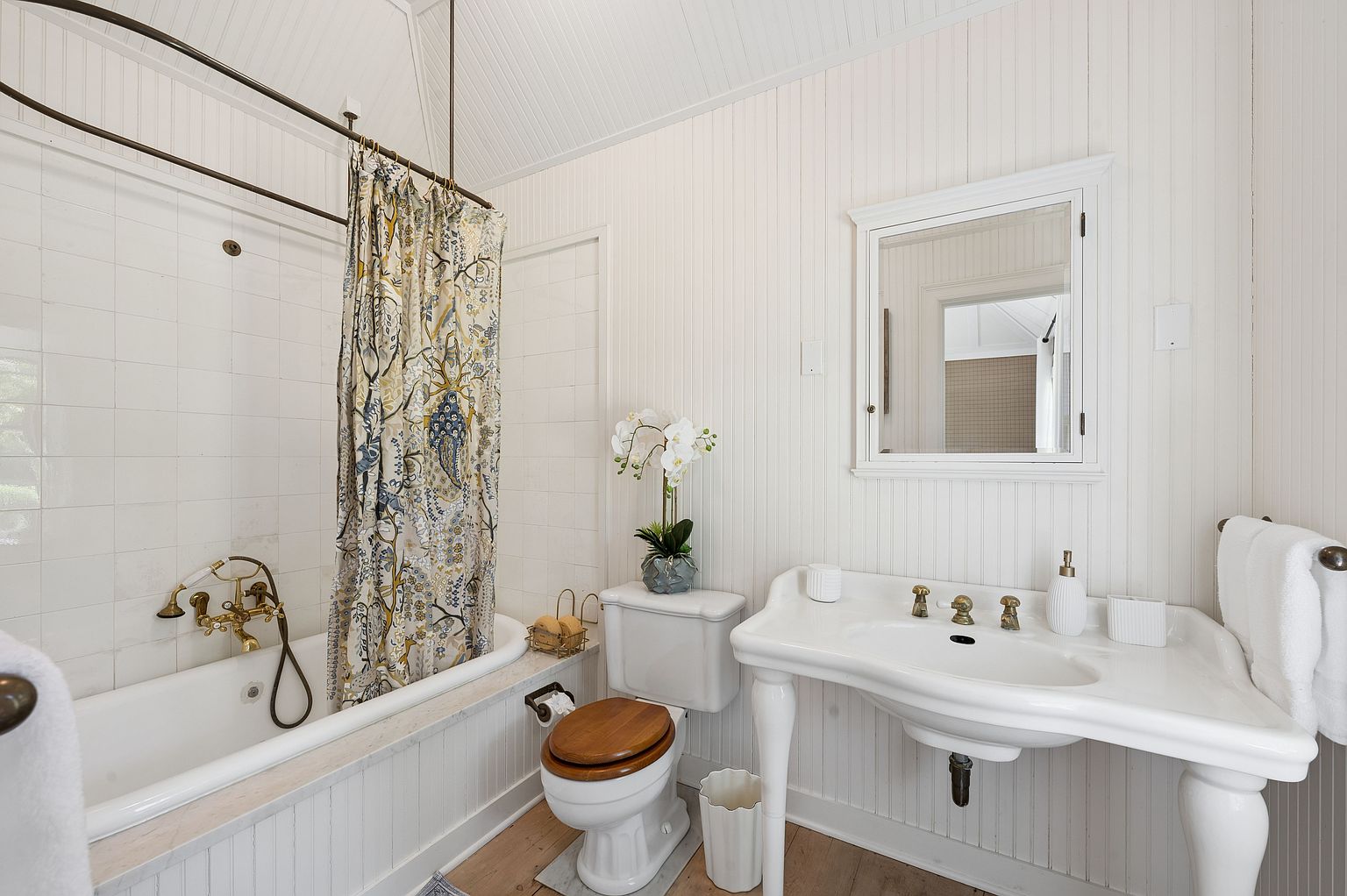 This bright, vintage-inspired bathroom features white beadboard paneling and a pedestal sink with brass-toned fixtures. A clawfoot-style tub is positioned under a shower curtain with a botanical print, complemented by a toilet with a wooden seat. The space is finished with light-toned wood flooring and a wall-mounted medicine cabinet, creating a clean and classic aesthetic.