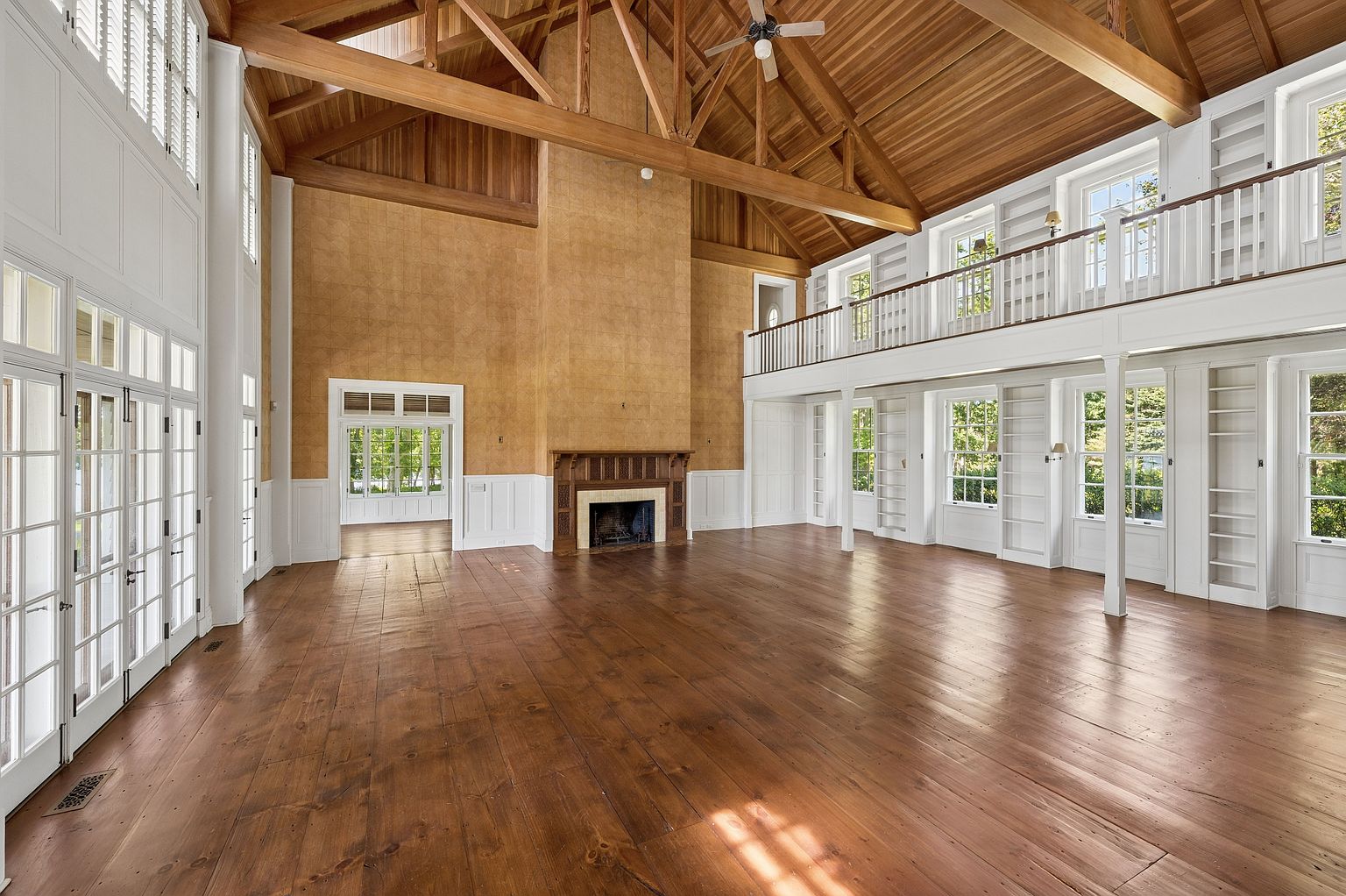 This expansive living room features a dramatic vaulted ceiling with exposed timber trusses and warm wood paneling. A central fireplace with a detailed wood mantel serves as a focal point against a textured, neutral-toned wall, while a mezzanine level with a white railing overlooks the main space. The room is finished with wide-plank wood flooring and numerous tall windows that flood the area with natural light, creating an airy and grand atmosphere.
