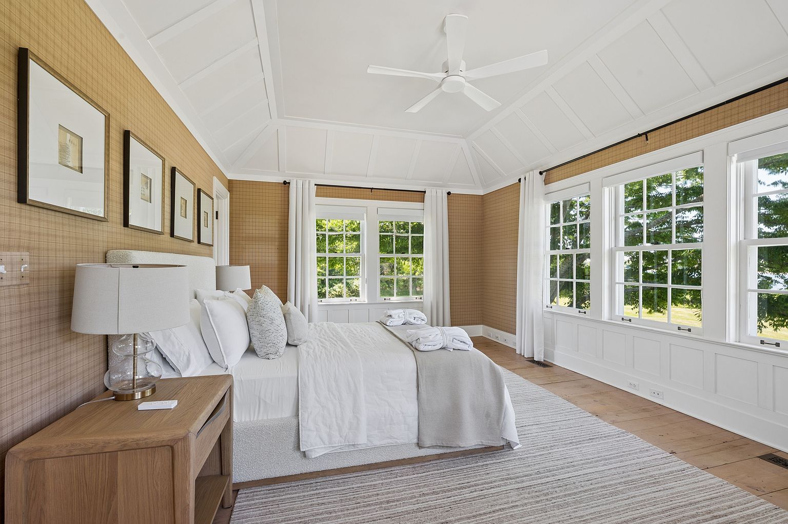 This bright and airy primary bedroom features a vaulted ceiling with a white fan and warm, textured wallpaper. A plush bed with neutral linens is centered against the wall, flanked by light wood nightstands and matching lamps. Large windows with white trim and curtains allow for ample natural light, while a striped area rug adds texture to the light wood flooring.