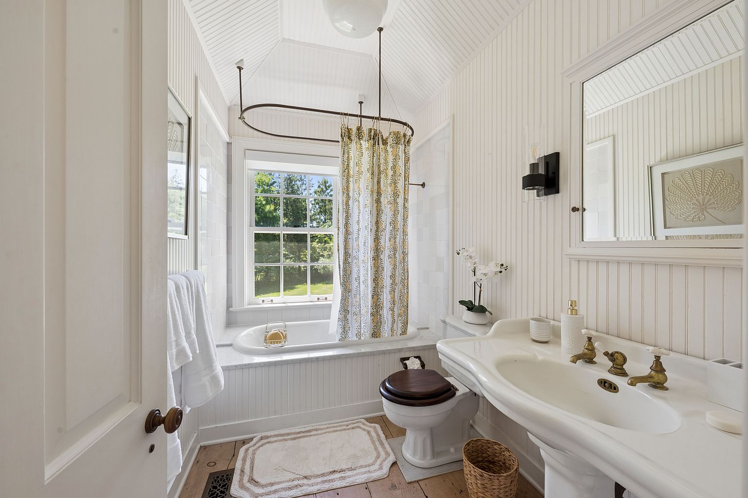 This bright, classic bathroom features white beadboard wainscoting and a vaulted ceiling, creating a clean and airy atmosphere. A pedestal sink with brass-toned fixtures sits beside a toilet with a dark wood seat, while a deep soaking tub is positioned beneath a window that overlooks a lush green yard. The space is finished with light wood flooring and a patterned shower curtain, offering a charming, traditional aesthetic.
