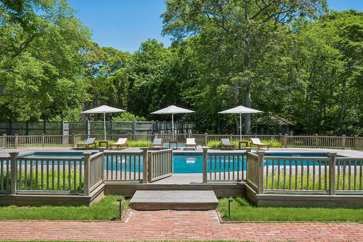 This serene backyard scene features a rectangular swimming pool surrounded by a wooden deck and a protective wooden fence. Several lounge chairs with white umbrellas are arranged neatly on the deck, creating an inviting space for relaxation. The area is framed by lush green trees, providing a private and tranquil atmosphere, with a brick walkway leading up to the pool gate.