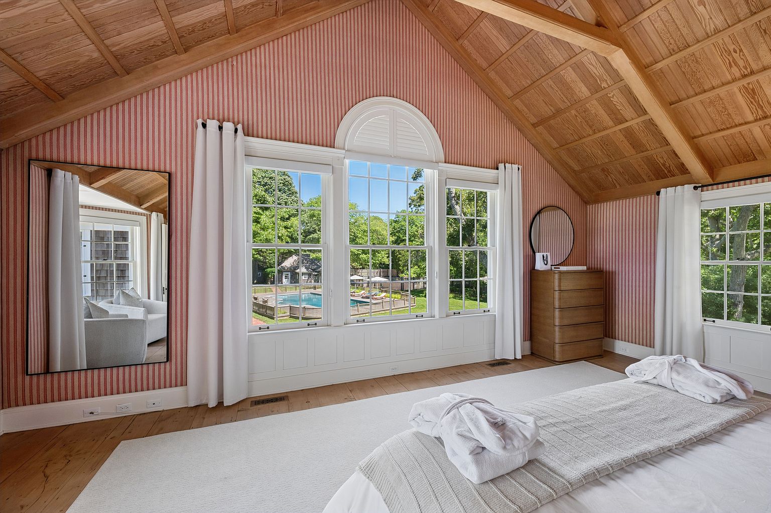 This bright and airy bedroom features a vaulted wood-paneled ceiling and distinctive pink-and-white striped wallpaper. A large central window assembly with an arched top provides a clear view of the backyard pool, while a large floor mirror and a wooden dresser add functional elegance. The room is finished with light wood flooring and a plush white area rug, creating a serene and inviting atmosphere.